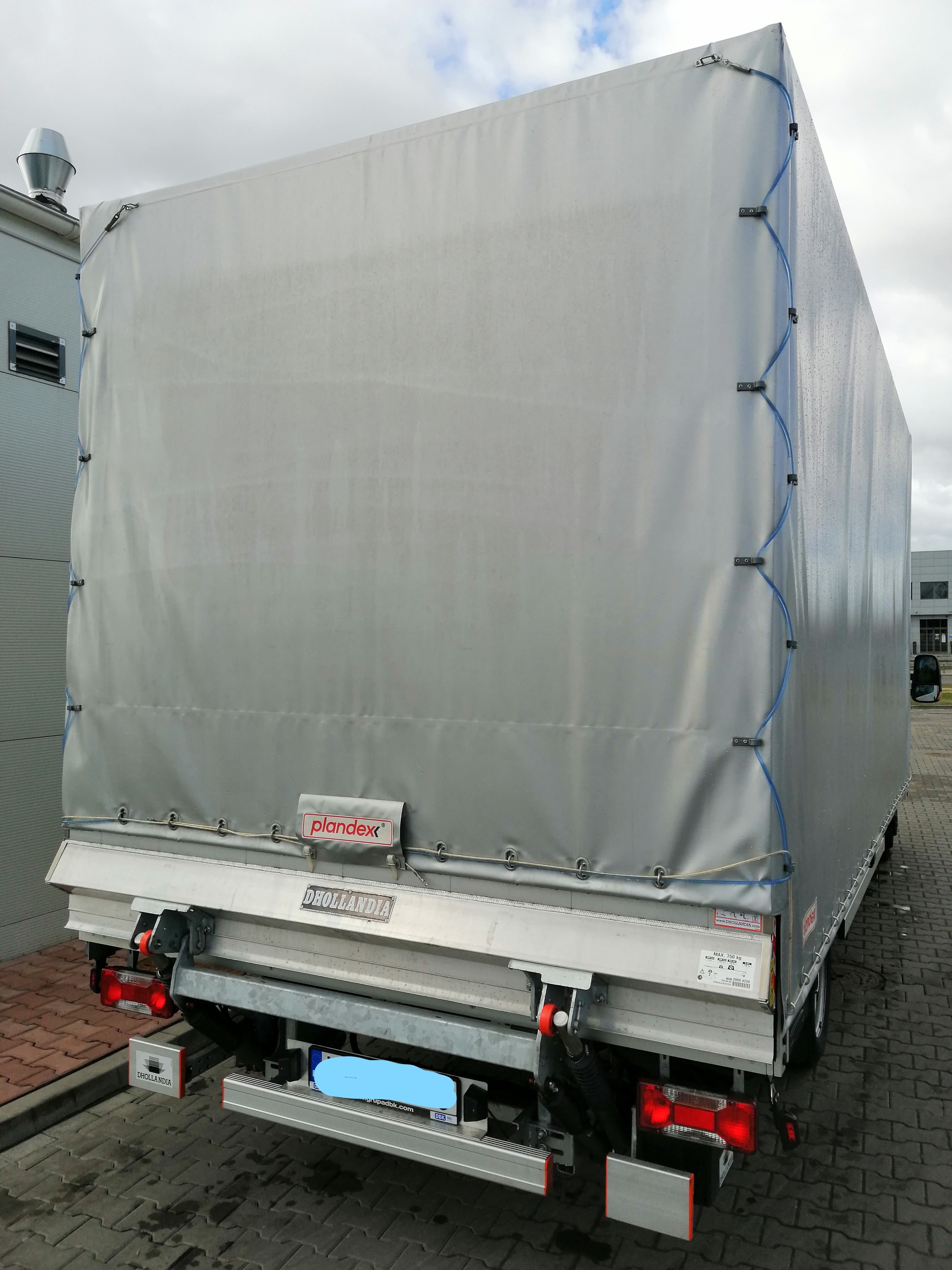 Tył samochodu dostawczego z plandeką, widoczne logo DHOLANDIA na burcie i plandex na plandece, oświetlenie tylne i elementy mocujące plandekę.