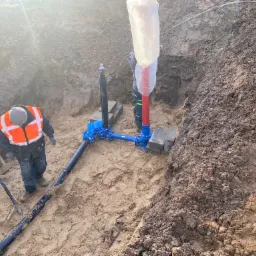 Instalacja rur w wykopie ziemnym z widocznymi złączkami i pracownikami w odblaskowych kamizelkach, zabezpieczenie rur w folii.