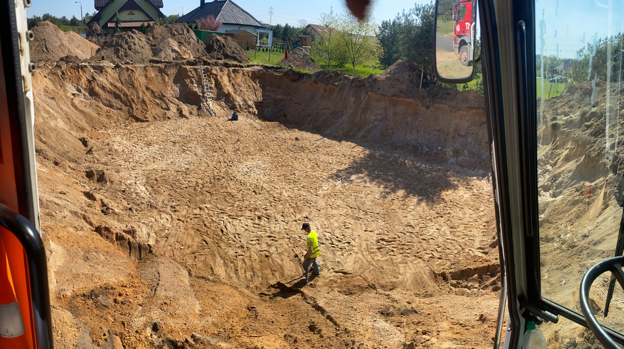 Głęboki wykop z piaszczystymi ścianami, widziany z kabiny maszyny budowlanej; pracownik w kamizelce odblaskowej na dnie wykopu, drabina oparta o jedną ze ścian, w tle zabudowa jednorodzinna...