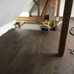 Układanie ciemnego parkietu w poddaszowym pokoju, widoczne narzędzia, fragment piły do cięcia, drewniane elementy konstrukcyjne dachu, doniczki z roślinami na tle białych ścian.