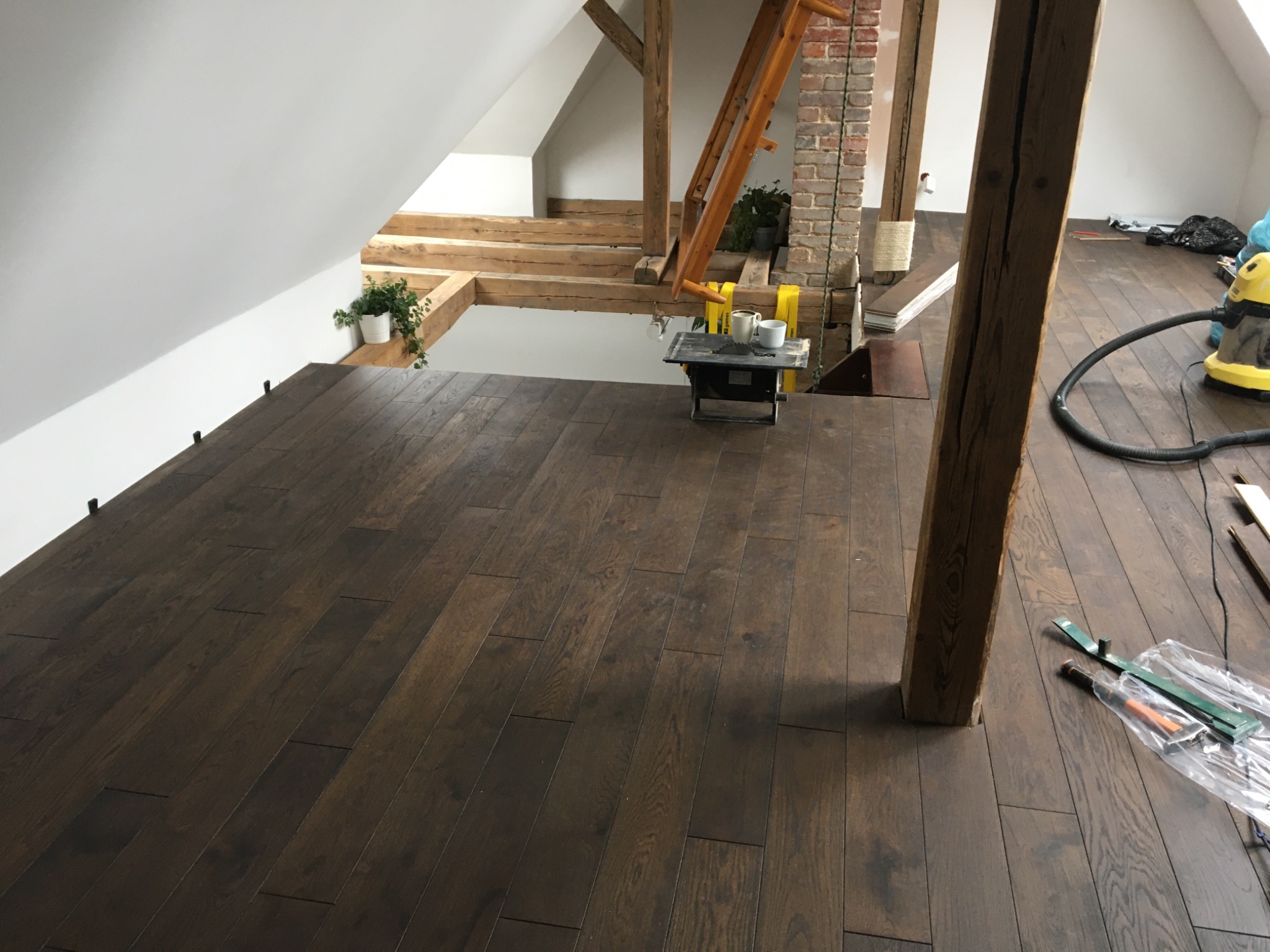 Układanie ciemnego parkietu w poddaszowym pokoju, widoczne narzędzia, fragment piły do cięcia, drewniane elementy konstrukcyjne dachu, doniczki z roślinami na tle białych ścian.