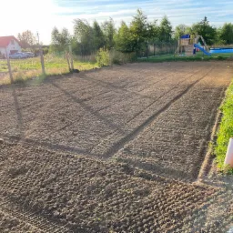 Przygotowany teren pod trawnik lub ogród, świeżo wyrównana ziemia z wyraźnymi śladami opon maszyny rolniczej, w tle dom jednorodzinny, ogrodzenie i plac zabaw z basenem.