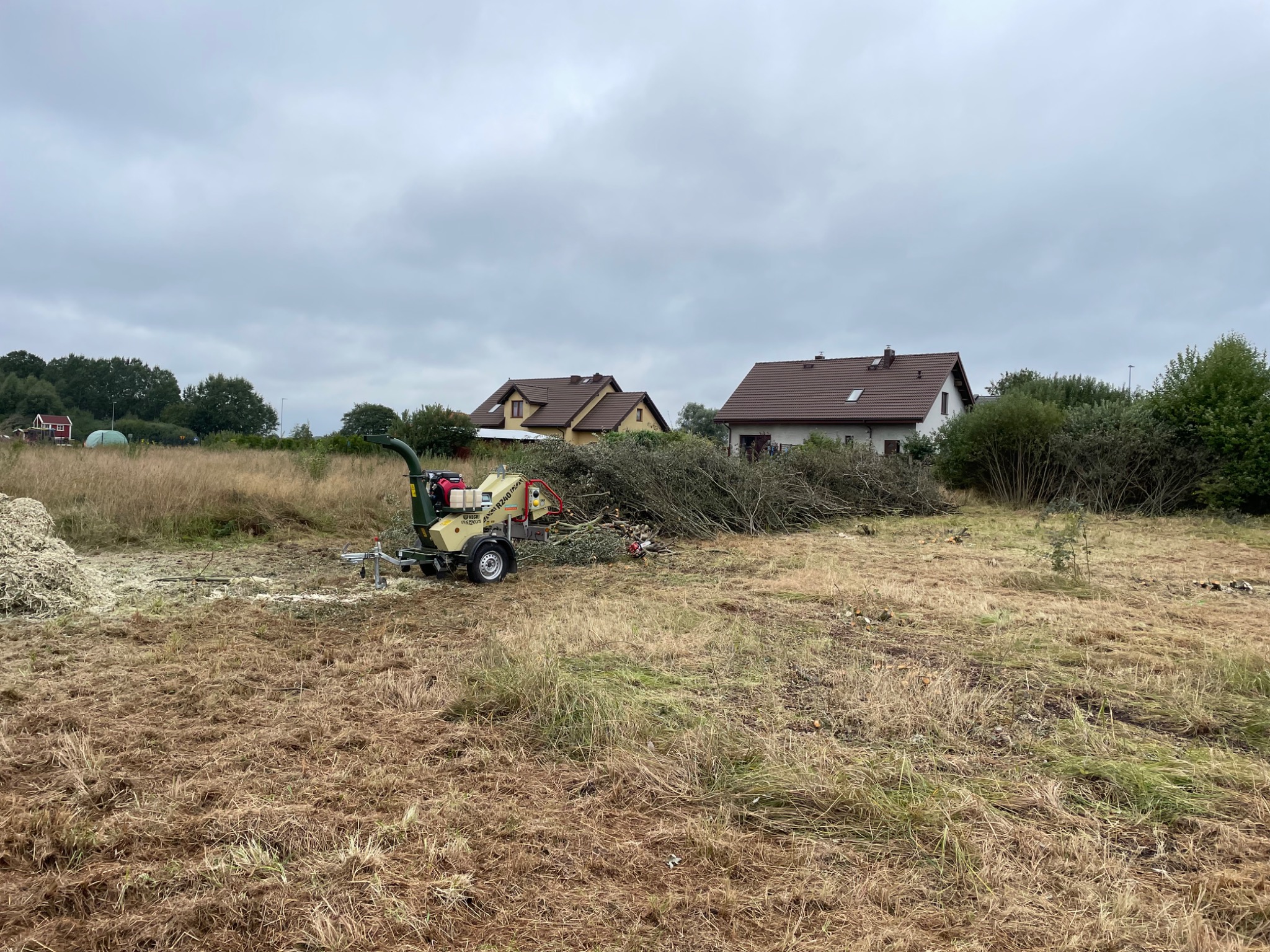 Profesjonalny rębak do gałęzi na działce, z pociętymi gałęziami i widocznymi domami w tle, pochmurny dzień.