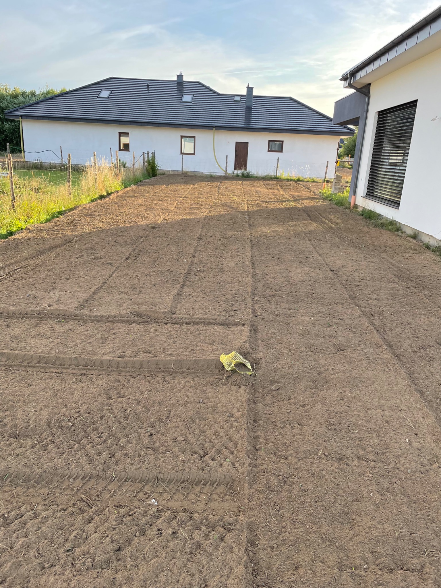 Przygotowany teren pod budowę domu jednorodzinnego, świeżo wyrównana ziemia z widocznymi śladami opon, worek foliowy leżący na ziemi, w tle biały dom z ciemnym dachem.