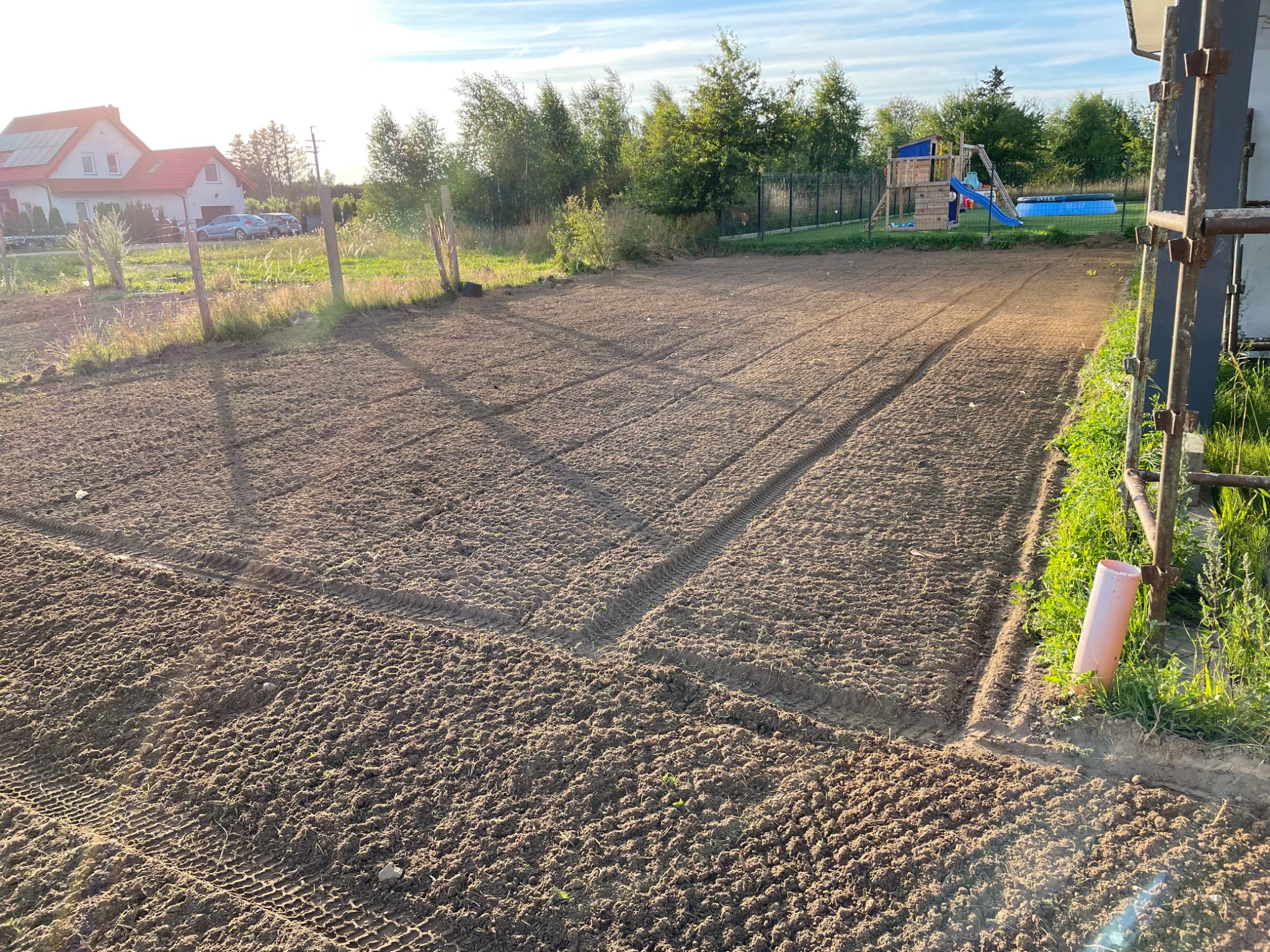 Przygotowany teren pod trawnik lub ogród, świeżo wyrównana ziemia z wyraźnymi śladami opon maszyny rolniczej, w tle dom jednorodzinny, ogrodzenie i plac zabaw z basenem.