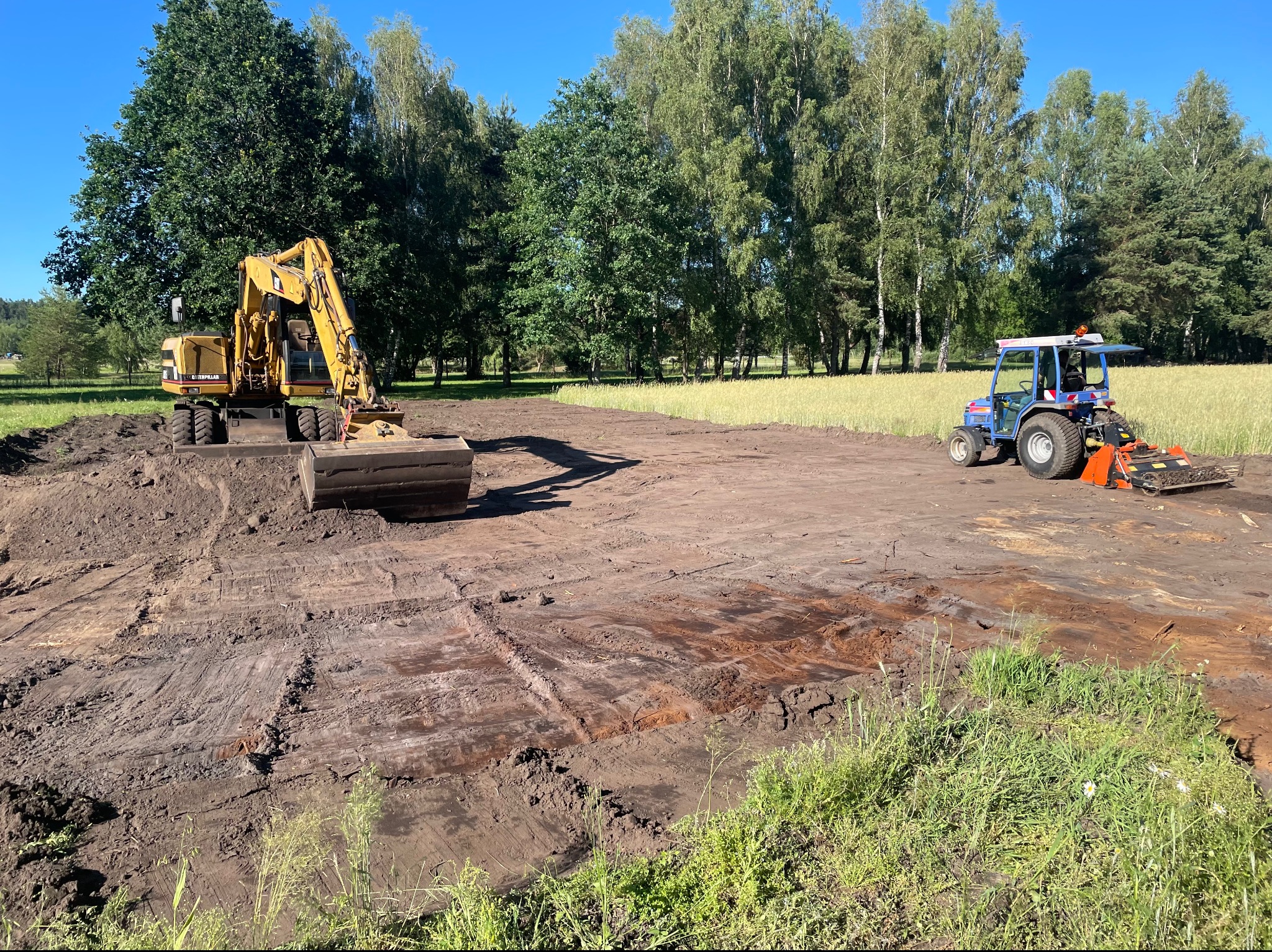 Żółta koparka Caterpillar i niebieski traktor z glebogryzarką na wyrównanym terenie, w tle zielone drzewa i pole zboża pod błękitnym niebem.
