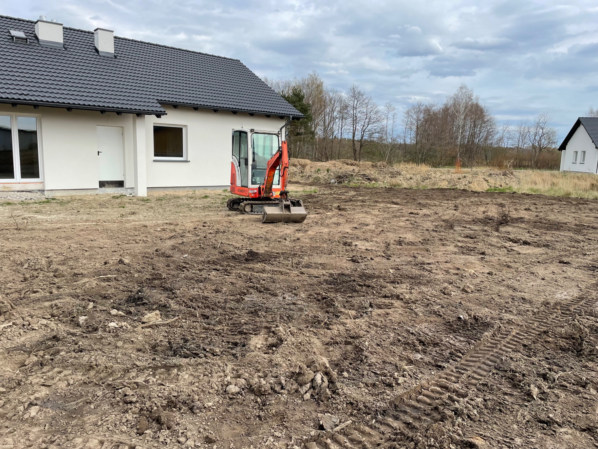 Czerwona mini koparka na nierównym, brązowym terenie budowy, w tle nowy dom z białymi ścianami i szarą dachówką oraz drzewa bez liści pod pochmurnym niebem.
