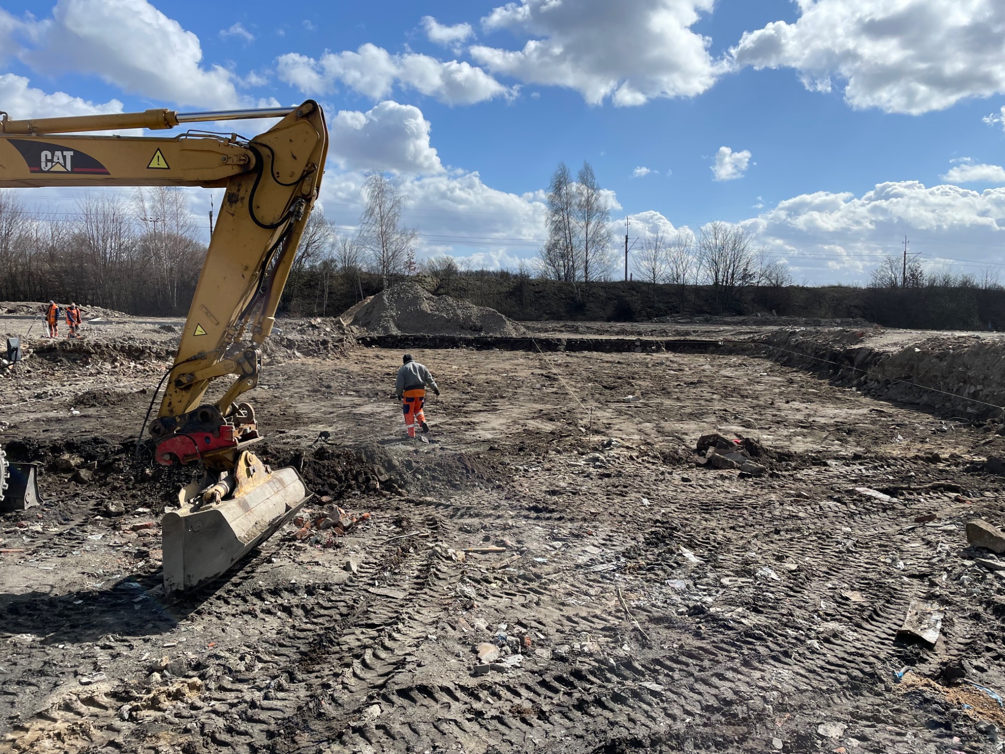 Żółta koparka CAT z łyżką w trakcie prac ziemnych na placu budowy pod błękitnym niebem z białymi chmurami, w tle pracownicy w pomarańczowych ubraniach.