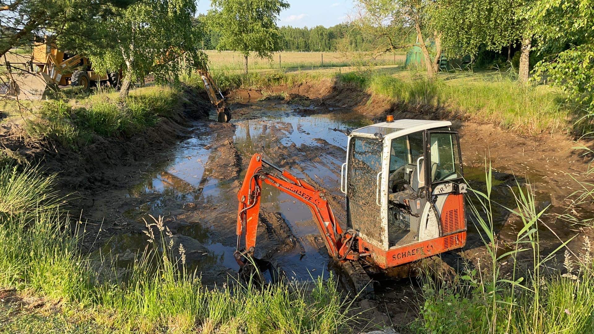 Pomarańczowa minikoparka Schaeff zanurzona w błotnistym wykopie stawu na tle zielonego krajobrazu z drzewami i łąką.