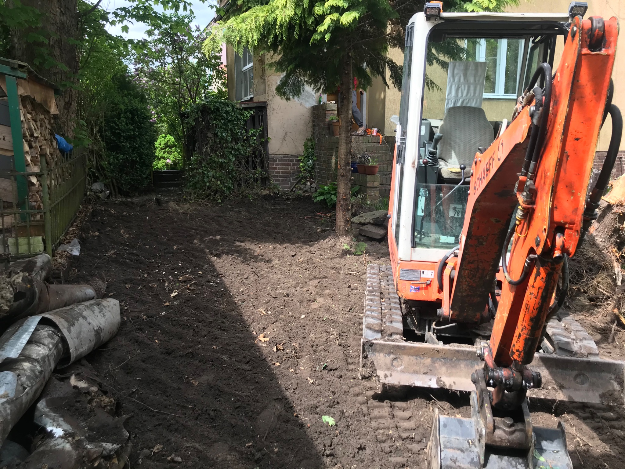 Pomarańczowa mini koparka na gąsienicach, przygotowująca teren pod budowę w ogrodzie, obok sterta starych rur