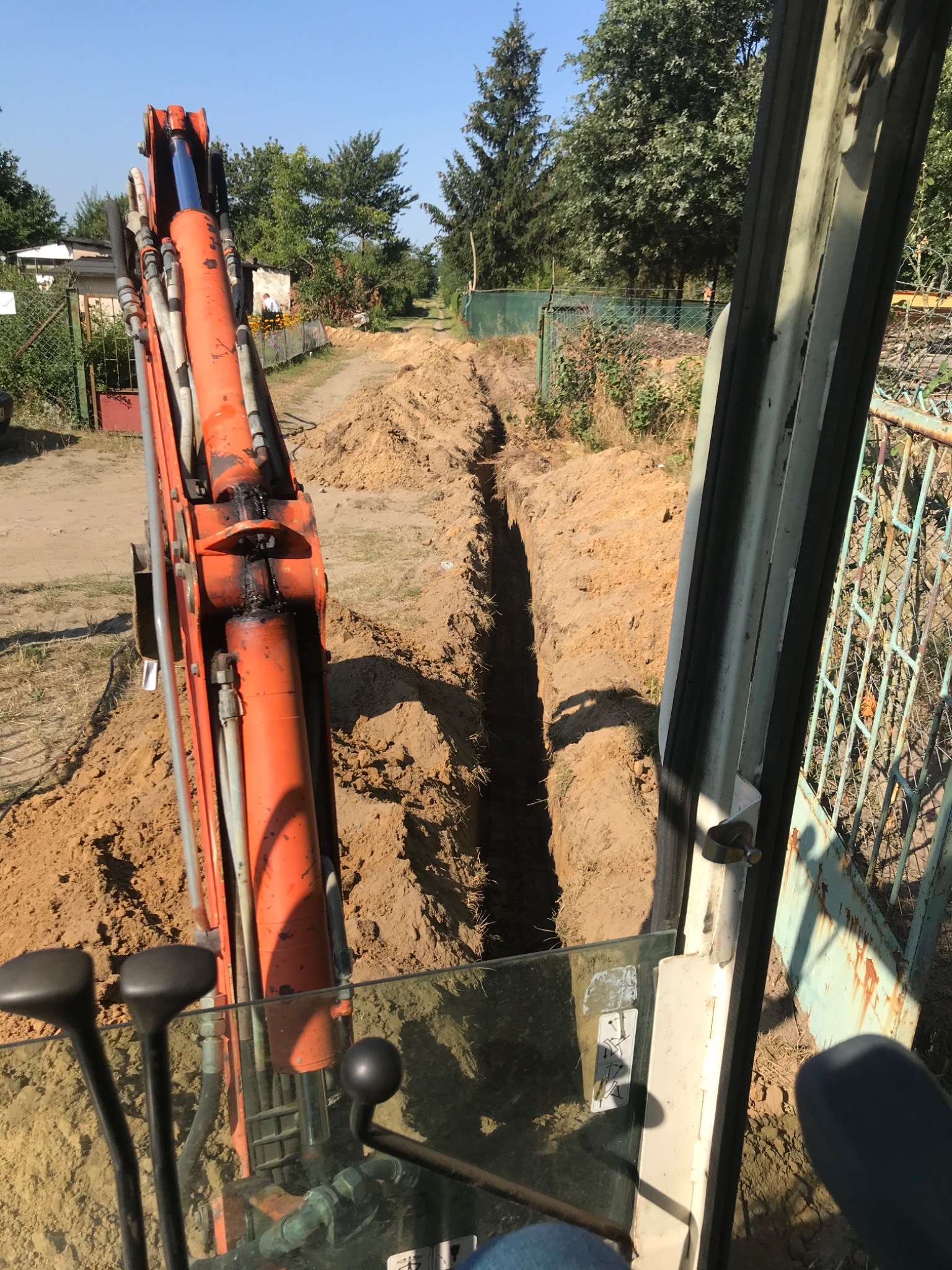 Widok z kabiny pomarańczowej koparki na wykopany wąski rów w ziemi, widoczny fragment ogrodzenia i drzewa w tle.
