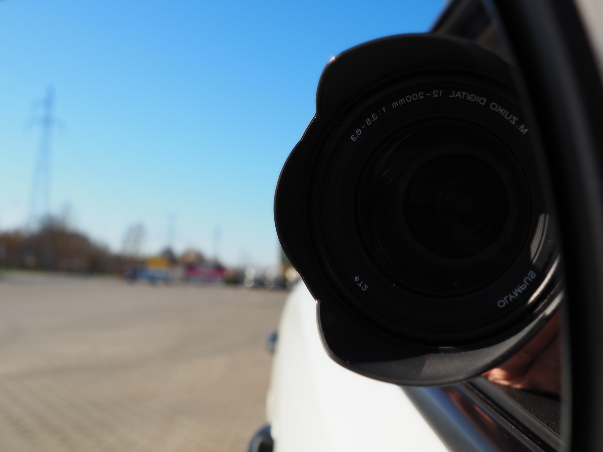 Obiektyw aparatu fotograficznego wystający z okna samochodu, w tle rozmyty parking i słupy wysokiego napięcia pod błękitnym niebem.