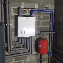 Zamknięta instalacja grzewcza w piwnicy z szarymi rurami, czerwonym zbiornikiem wyrównawczym i szarą ścianą z betonowych bloków.
