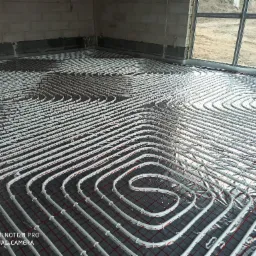 Białe rury ogrzewania podłogowego ułożone spiralnie na szarej folii z czerwoną siatką w surowym wnętrzu, widoczne okno i betonowe ściany.