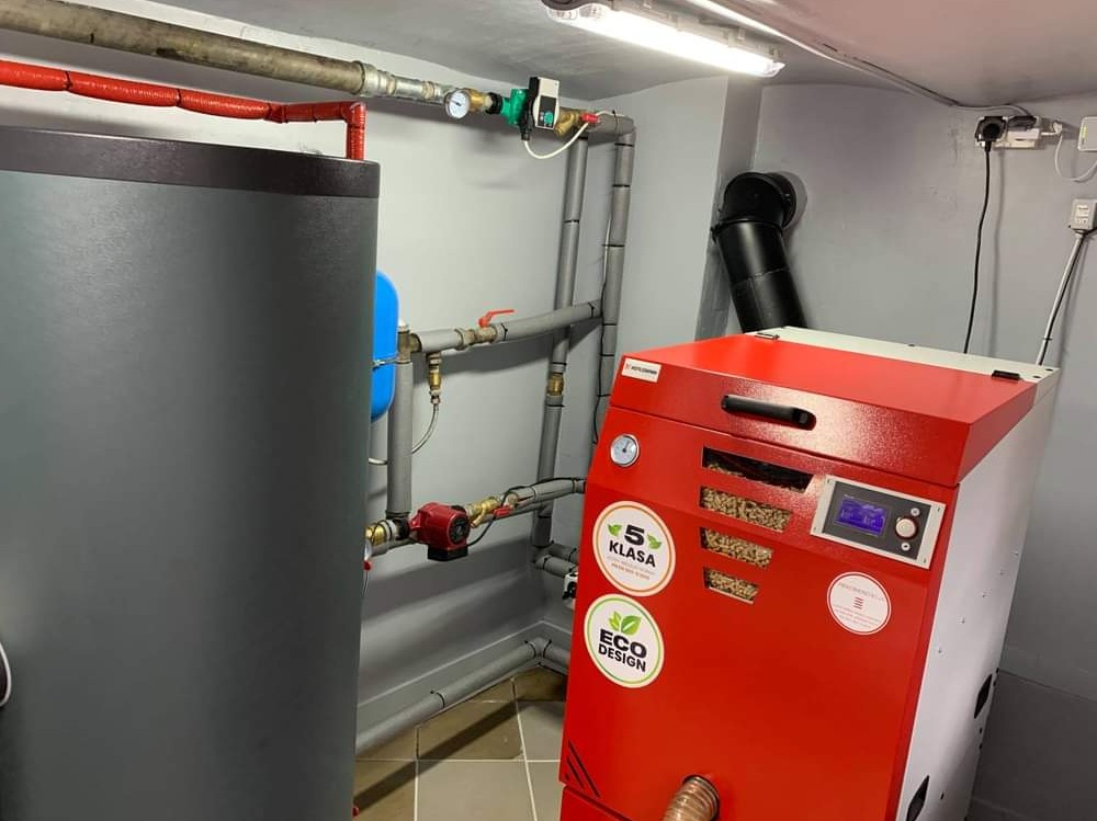Nowoczesna kotłownia z kotłem na pellet w kolorze czerwonym z naklejkami '5 Klasa' i 'Eco Design', obok szary zbiornik na wodę, instalacja grzewcza z rurami i zaworami.