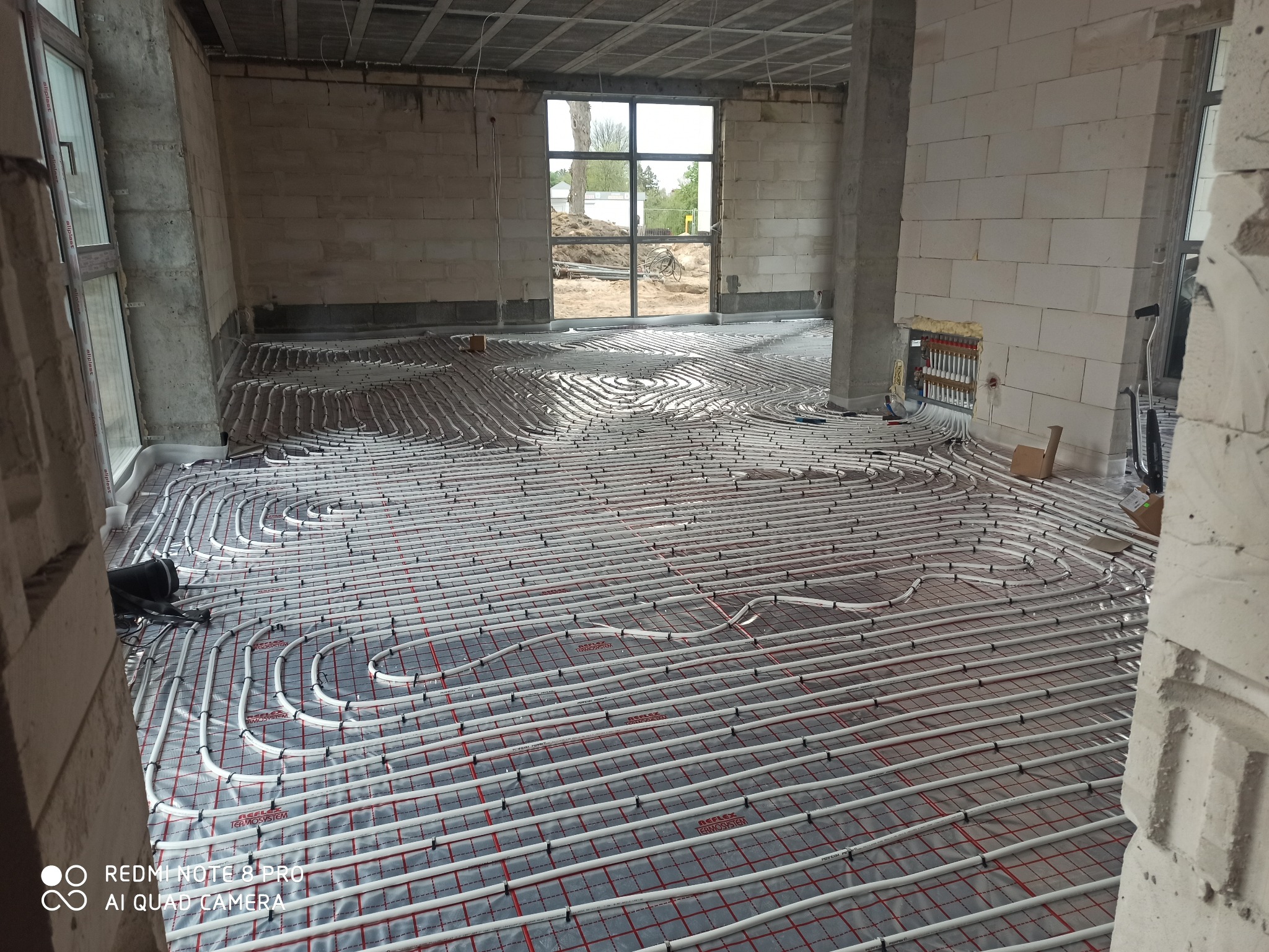 Widok na rozłożone rury ogrzewania podłogowego na folii z siatką w surowym wnętrzu budynku w trakcie budowy, z widoczną rozdzielnicą na ścianie i oknem w tle.