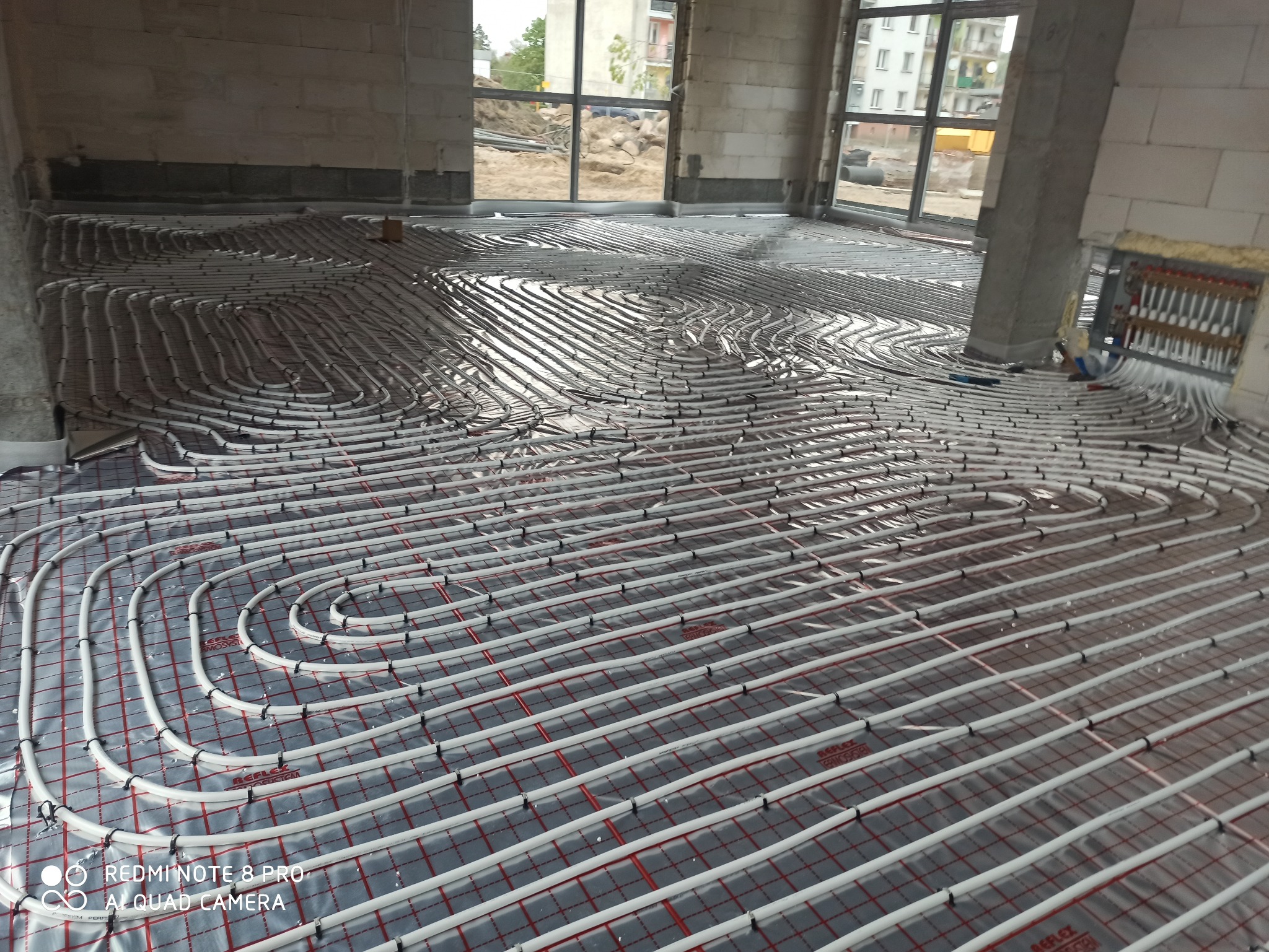Rozłożona instalacja ogrzewania podłogowego z rurami na folii z siatką, widoczny rozdzielacz i surowy stan budynku.