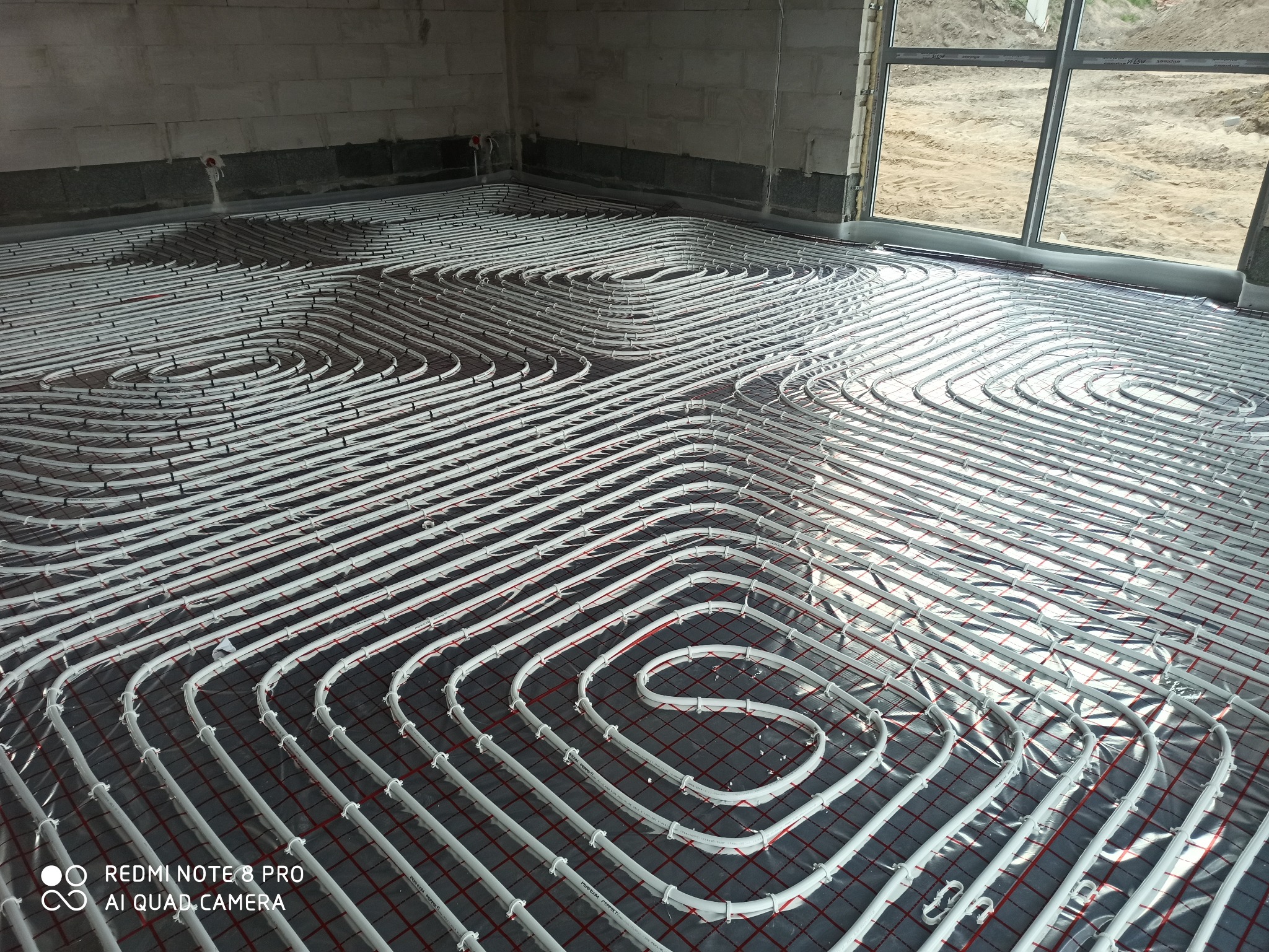 Białe rury ogrzewania podłogowego ułożone spiralnie na szarej folii z czerwoną siatką w surowym wnętrzu, widoczne okno i betonowe ściany.
