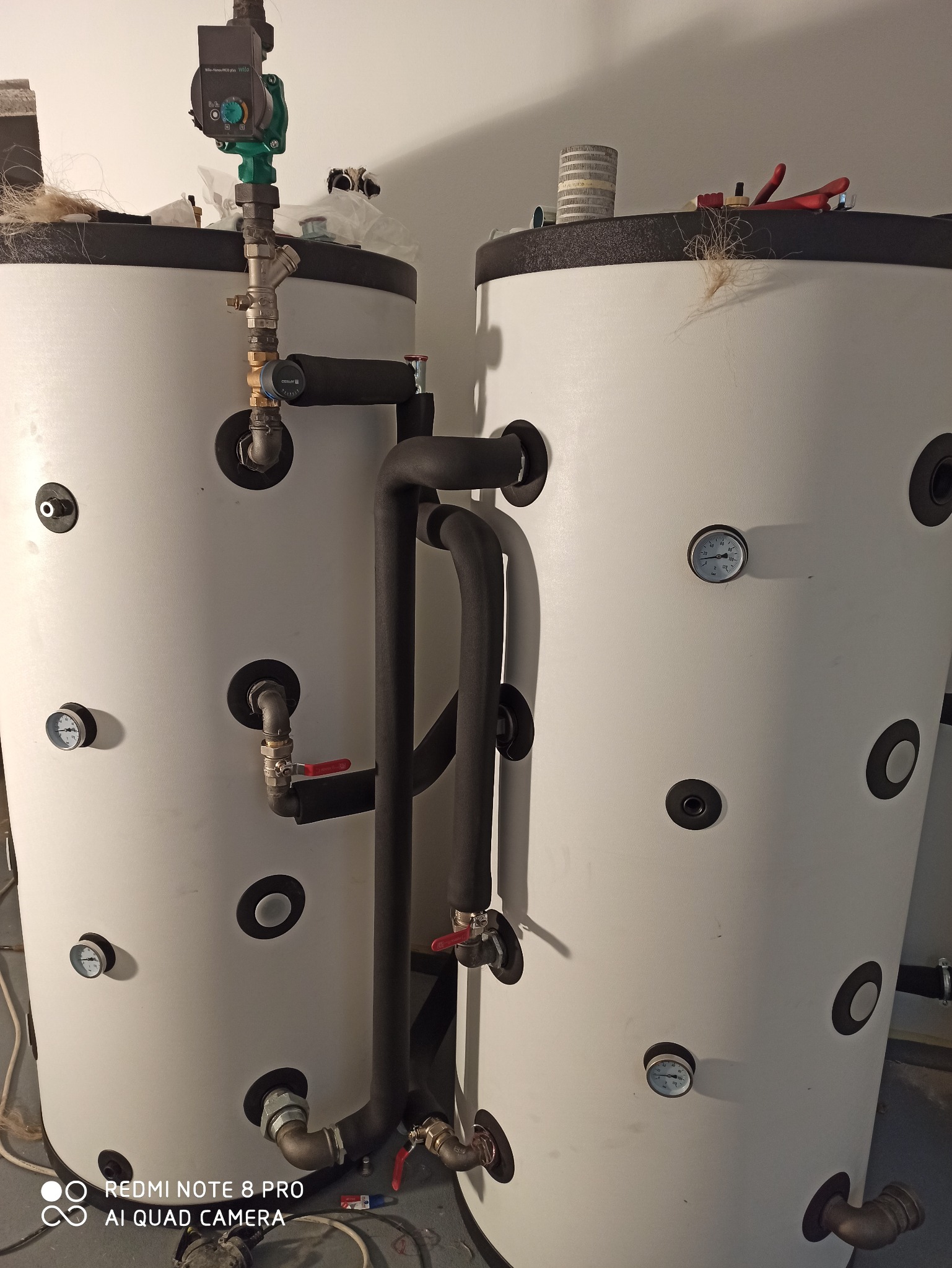 Dwa białe zbiorniki buforowe z czarnymi rurami i zaworami, w tle biała ściana, widoczne mierniki temperatury i narzędzia na górze zbiornika.