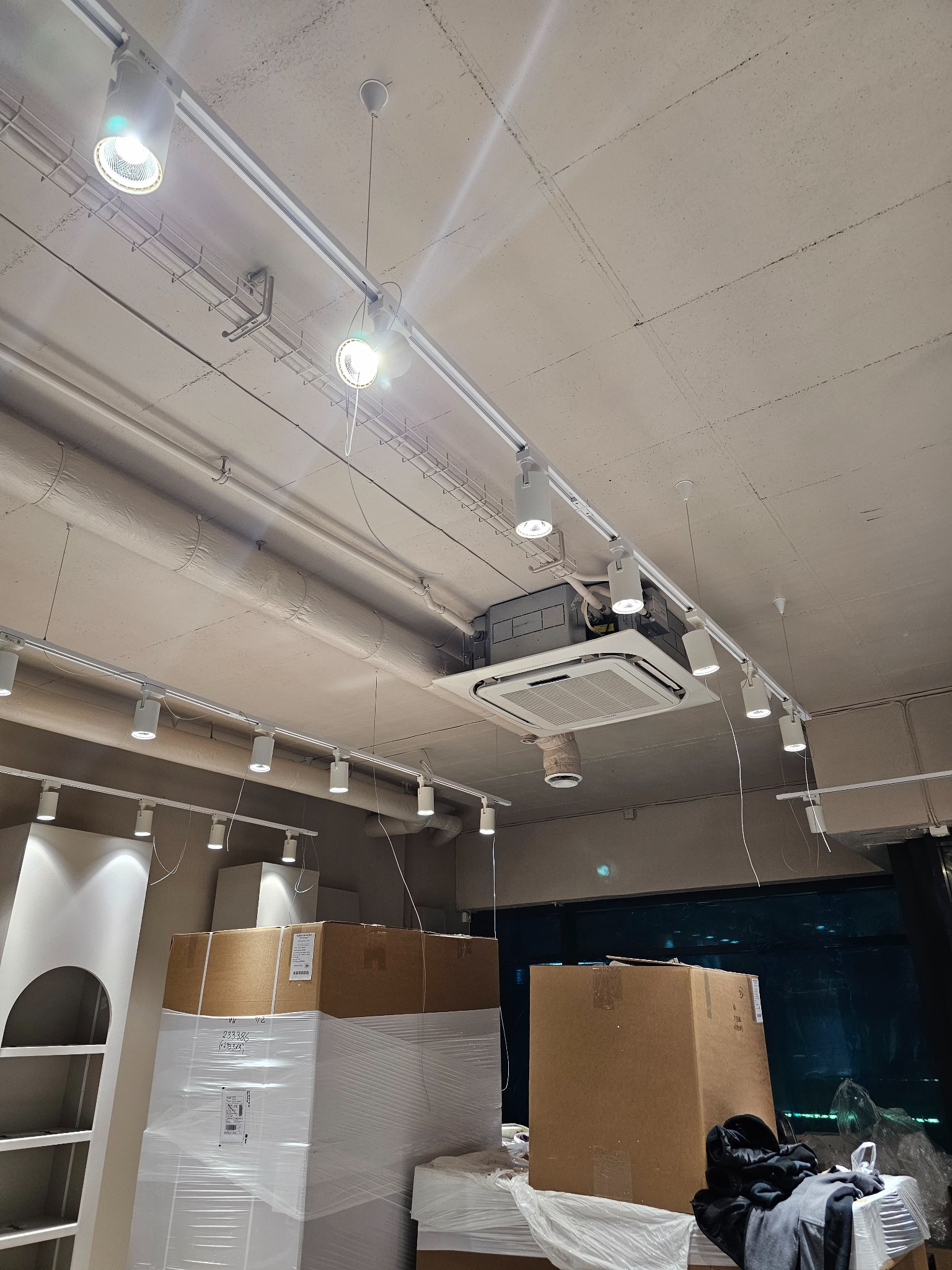 Sufitowe oświetlenie szynowe z zamontowaną kasetą klimatyzacyjną w lokalu komercyjnym, widoczne kartony z zapakowanymi elementami wyposażenia.