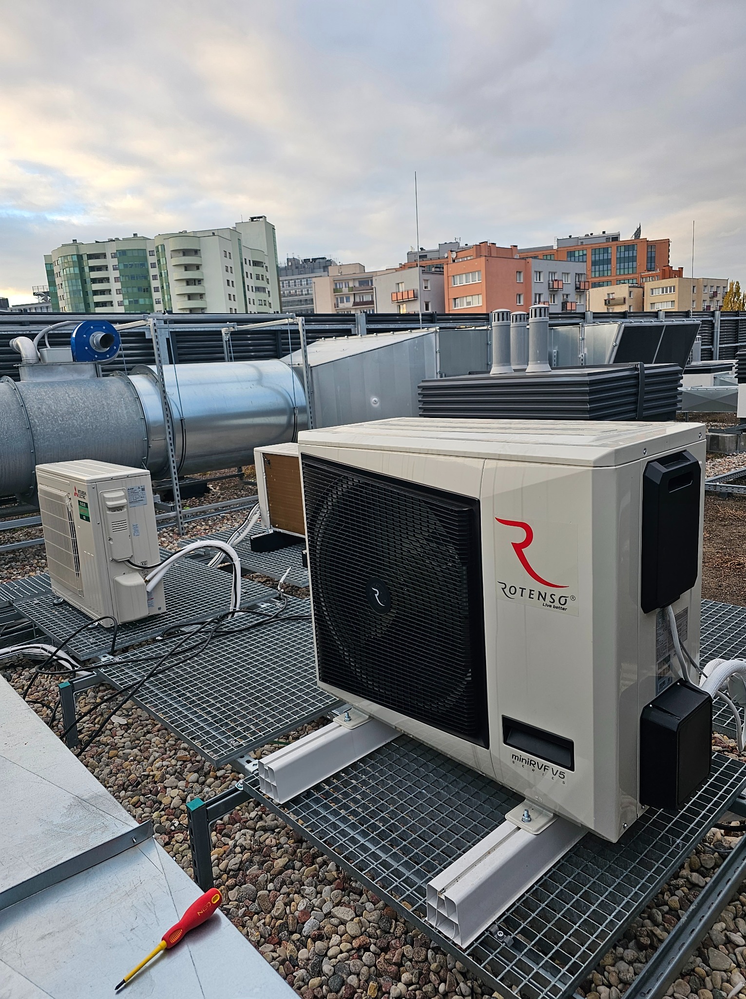 Instalacja Rotenso miniVRF V5 na dachu budynku, widoczne przewody, elementy wentylacyjne i narzędzie - śrubokręt leżący na dachu. W tle zabudowa miejska pod pochmurnym niebem.