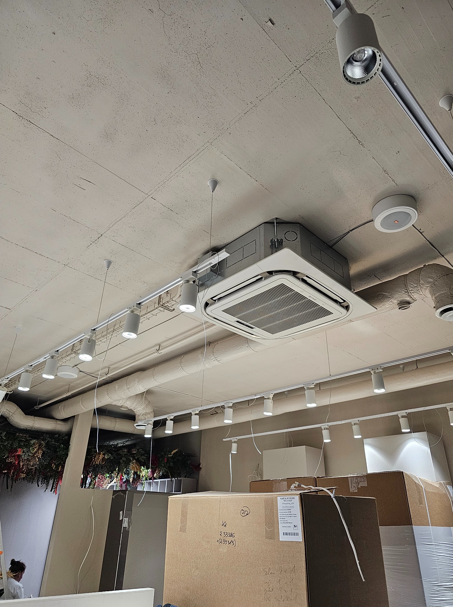 Kasetonowa jednostka klimatyzacyjna zamontowana pod betonowym sufitem, widoczne rury wentylacyjne i oświetlenie szynowe, na pierwszym planie kartony z towarem.