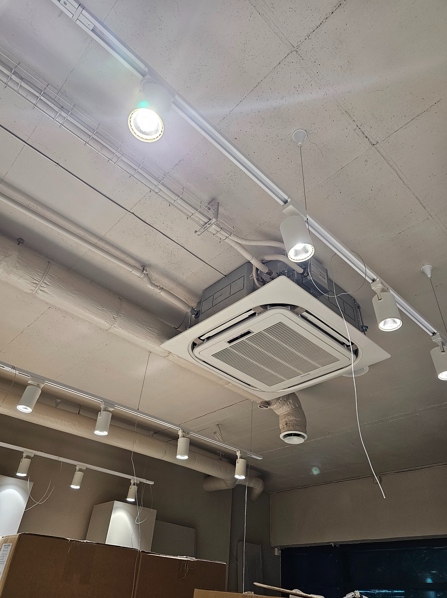 Czterokierunkowa klimatyzacja kasetonowa zamontowana w suficie biura z widocznymi przewodami i oświetleniem szynowym.