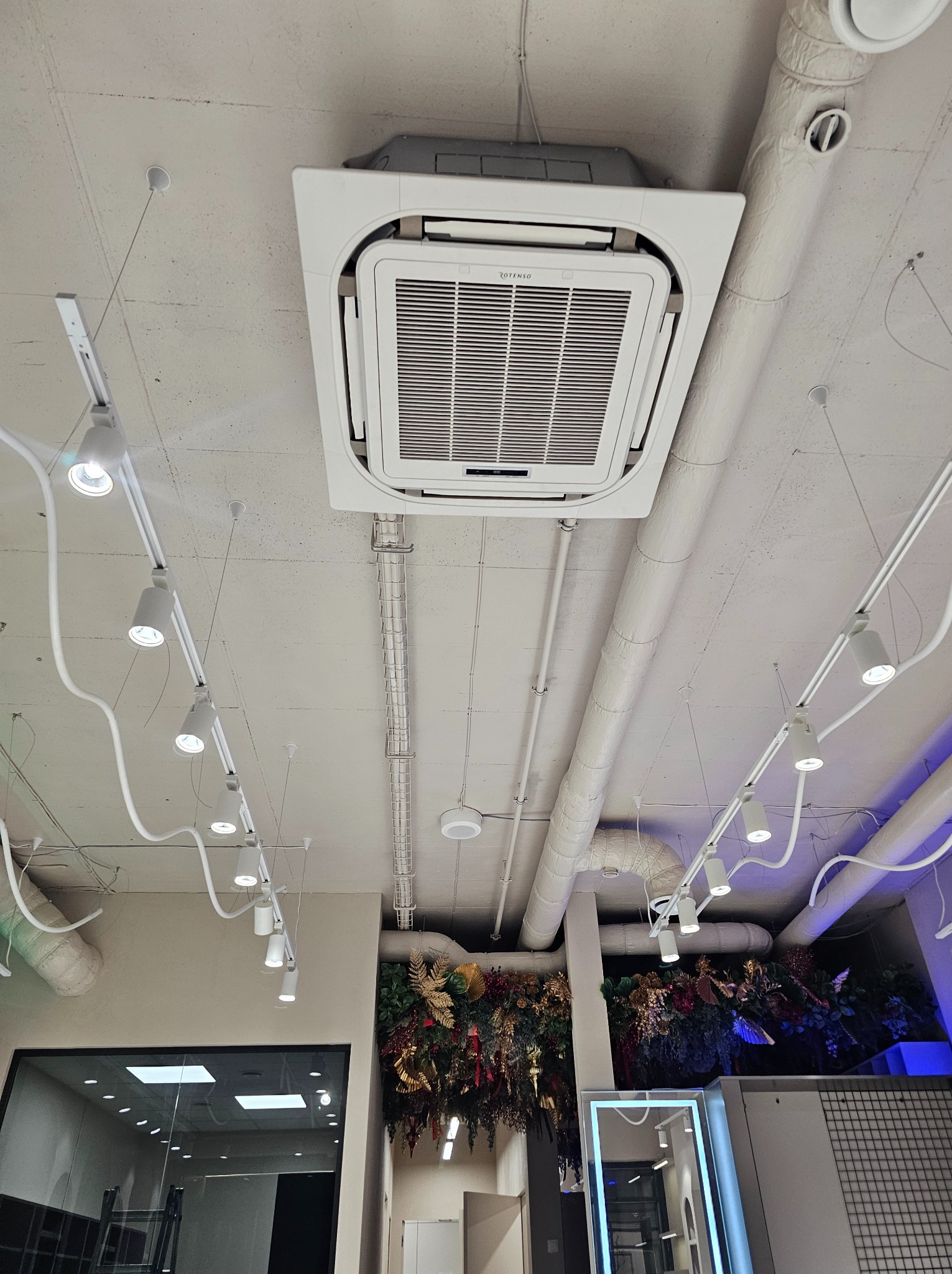 Sufit biura z widoczną kasetą klimatyzacyjną Rotenso, rurami wentylacyjnymi i oświetleniem punktowym na szynoprzewodach.