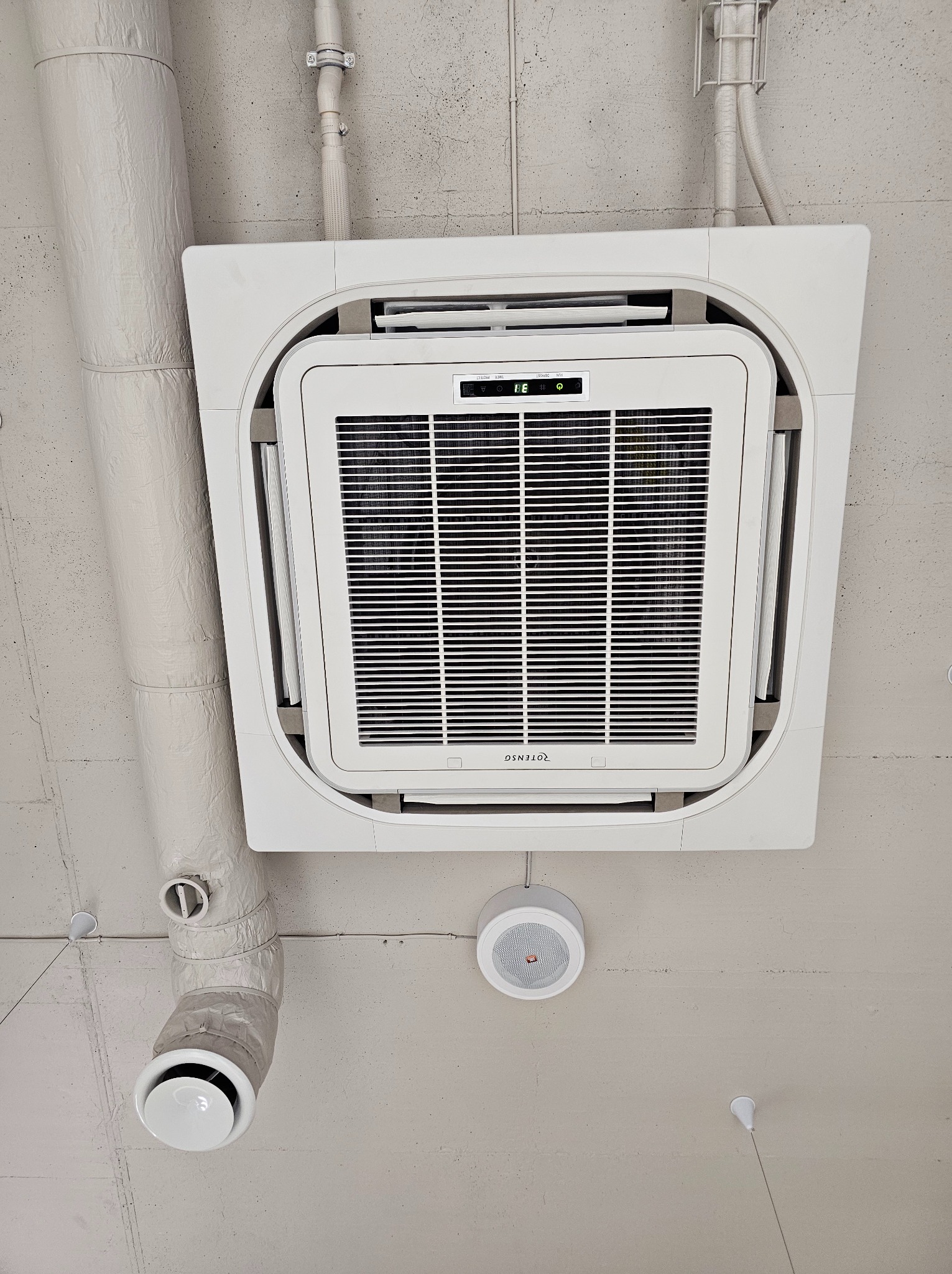 Kasetonowa jednostka klimatyzacyjna z widocznym wyświetlaczem temperatury, zamontowana na betonowym suficie, z widocznymi elementami instalacji wentylacyjnej i głośnikiem sufitowym.