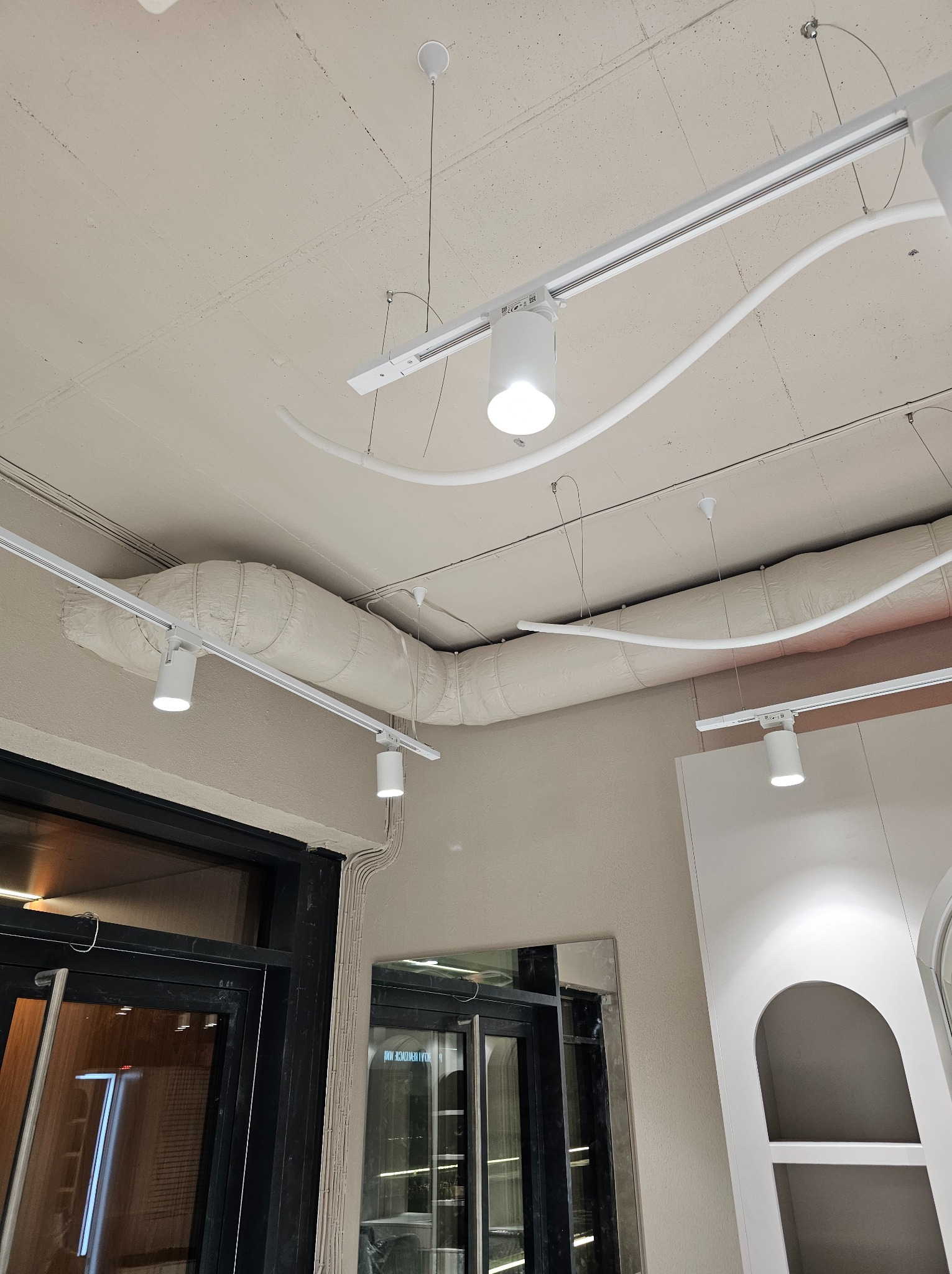 Widok na sufit z widocznymi elementami instalacji: rurami wentylacyjnymi w izolacji, szynami oświetleniowymi z reflektorami punktowymi oraz przewodami elektrycznymi.