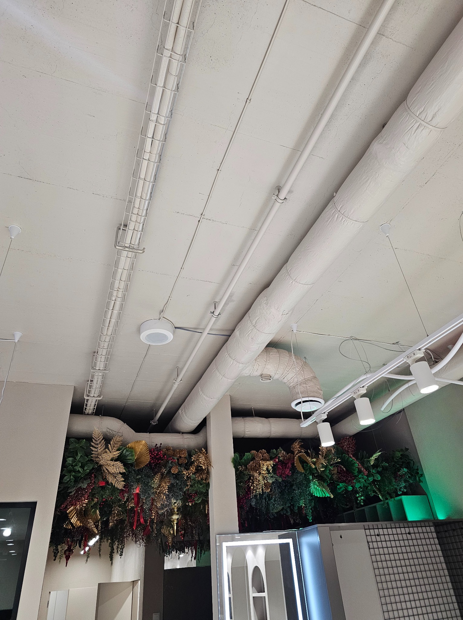 Widok na sufit w biurze z widocznymi elementami instalacji wentylacyjnych, w tym kanałami owiniętymi izolacją, metalowymi korytkami kablowymi i oświetleniem punktowym. Dodatkowo widać dekoracje...