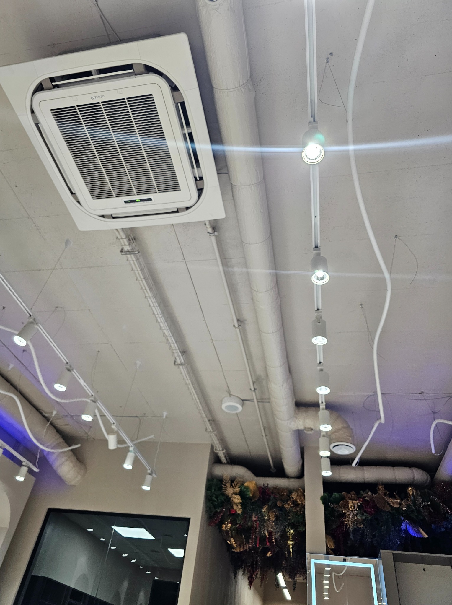 Kasetonowa jednostka klimatyzacyjna zamontowana na suficie podwieszanym w pomieszczeniu komercyjnym, widoczne instalacje elektryczne i oświetleniowe.