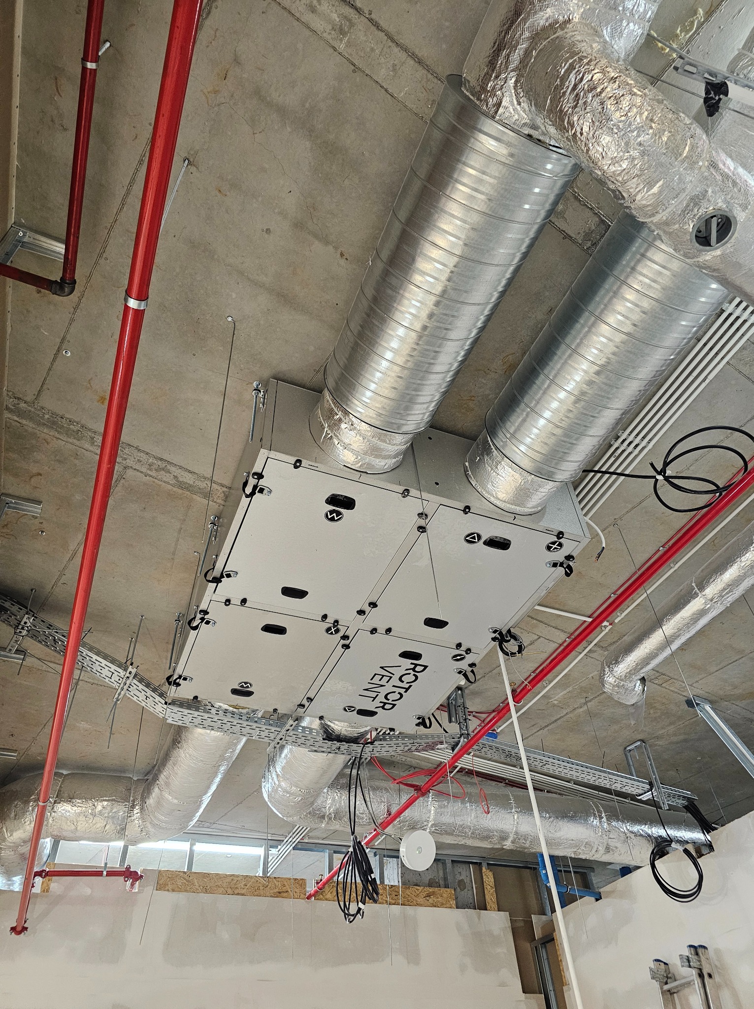 Widok instalacji sufitowej z centralą wentylacyjną Rotor Vent, rurami spiro owiniętymi folią aluminiową, czerwonymi rurami instalacji przeciwpożarowej i korytami kablowymi pod betonowym stropem.
