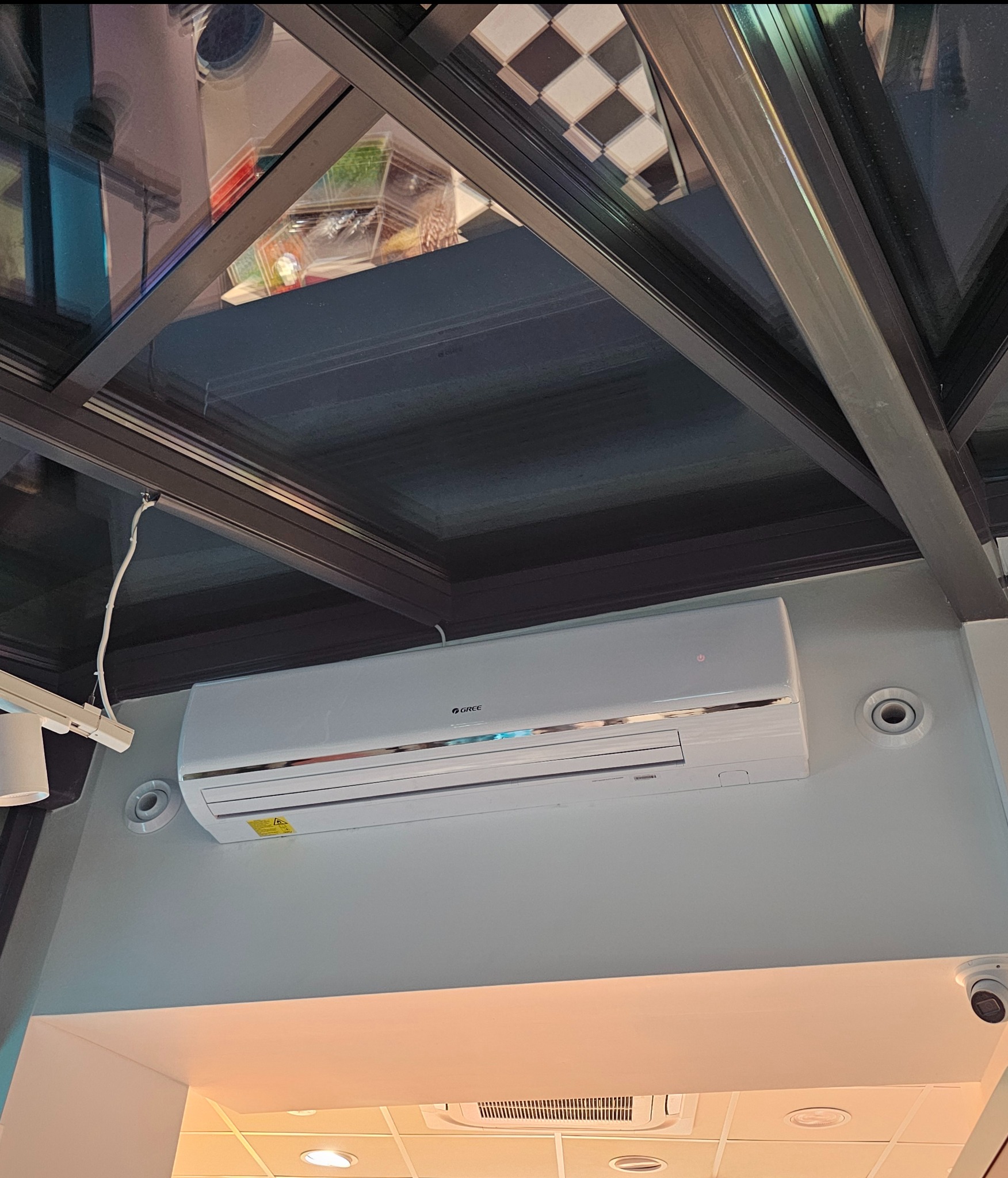 Klimatyzator marki Gree zamontowany pod przeszklonym sufitem z czarną ramą, widoczny także fragment sufitu z kratką i oświetleniem oraz kamera monitoringu