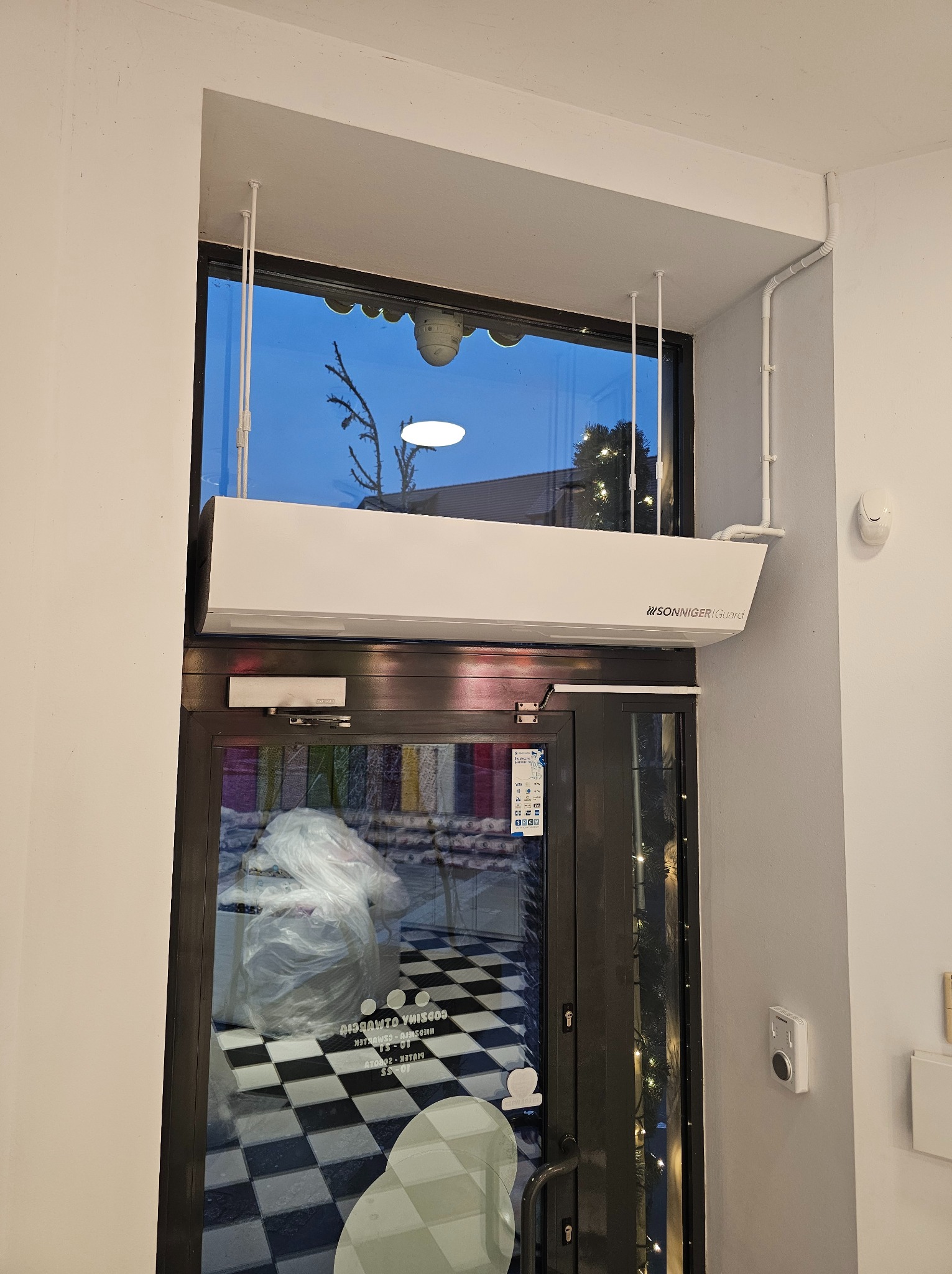 Biała kurtyna powietrzna marki Sonniger Guard zamontowana nad wejściem do sklepu z czarno-białą podłogą w kratkę, widoczna przez szybę