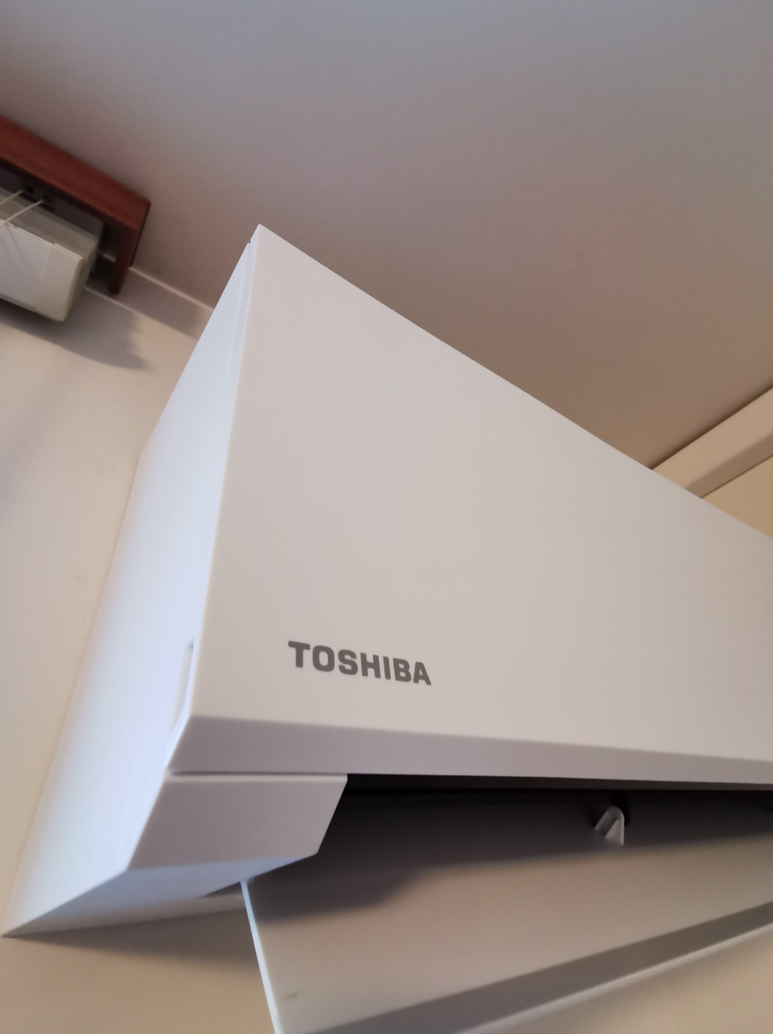 Biała jednostka wewnętrzna klimatyzatora Toshiba zamontowana pod sufitem, widoczna z perspektywy dolnej, z fragmentem kratki wentylacyjnej w tle.