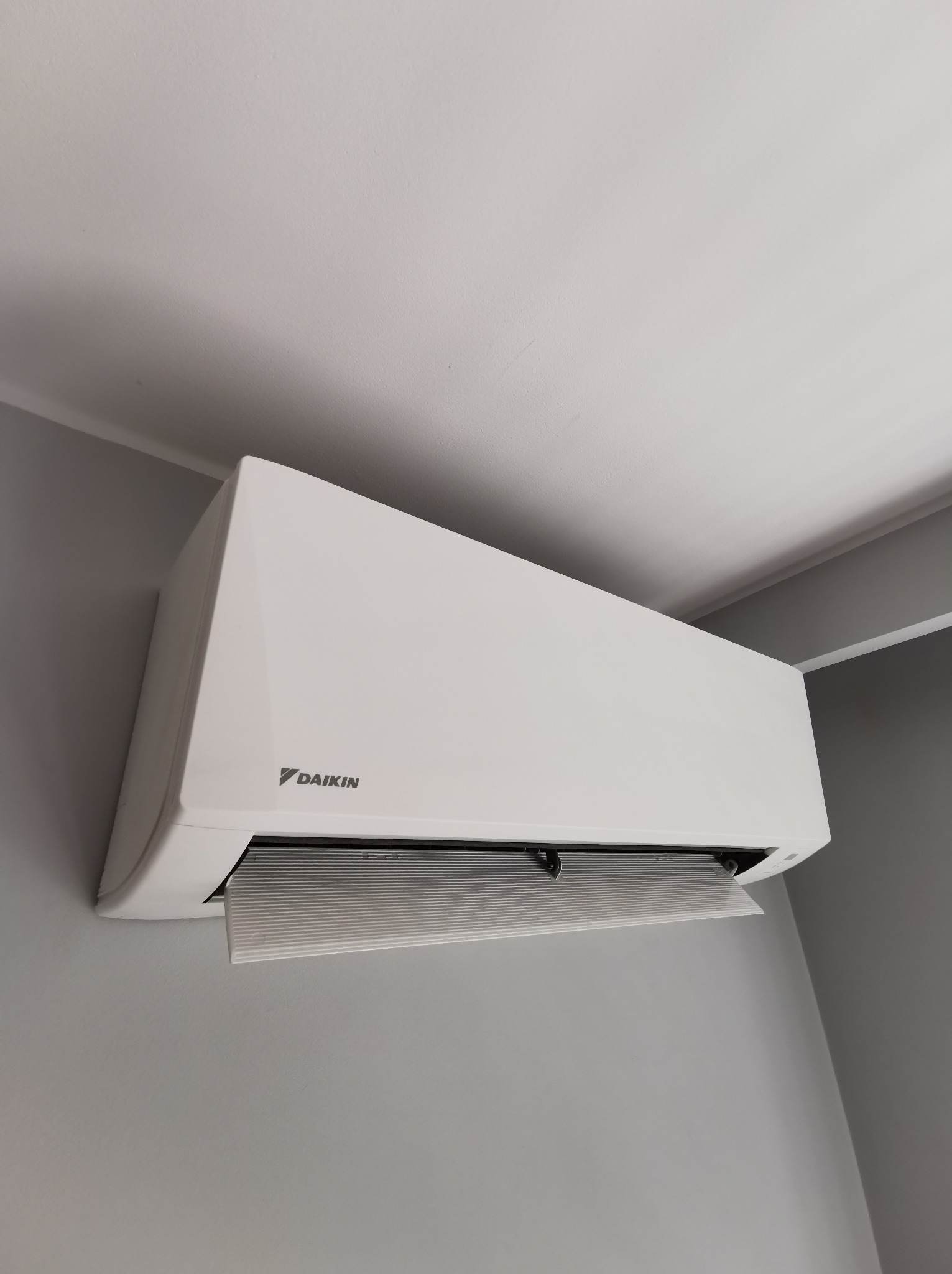 Biała klimatyzacja Daikin zamontowana na szarej ścianie, widok z dołu, z otwartą kratką wentylacyjną.
