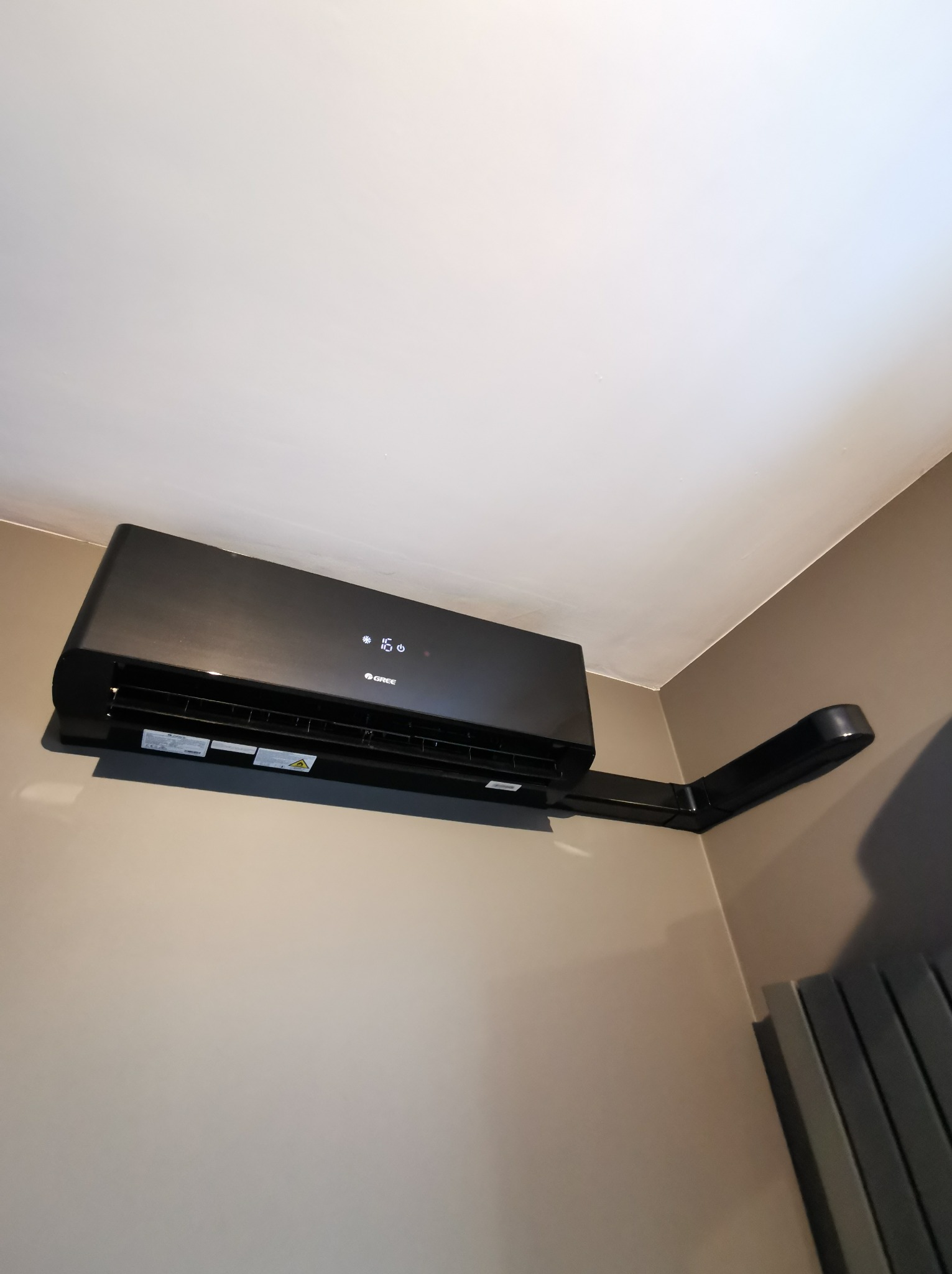 Czarny klimatyzator marki Gree zamontowany na ścianie w pomieszczeniu o beżowych ścianach, widoczny przewód odprowadzający skropliny.