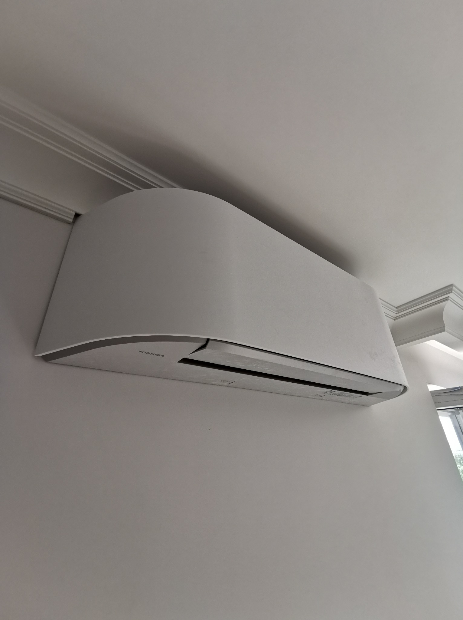 Biała jednostka klimatyzacyjna Toshiba zamontowana na ścianie w jasnym pomieszczeniu, widoczny fragment okna i ozdobny gzyms sufitowy.