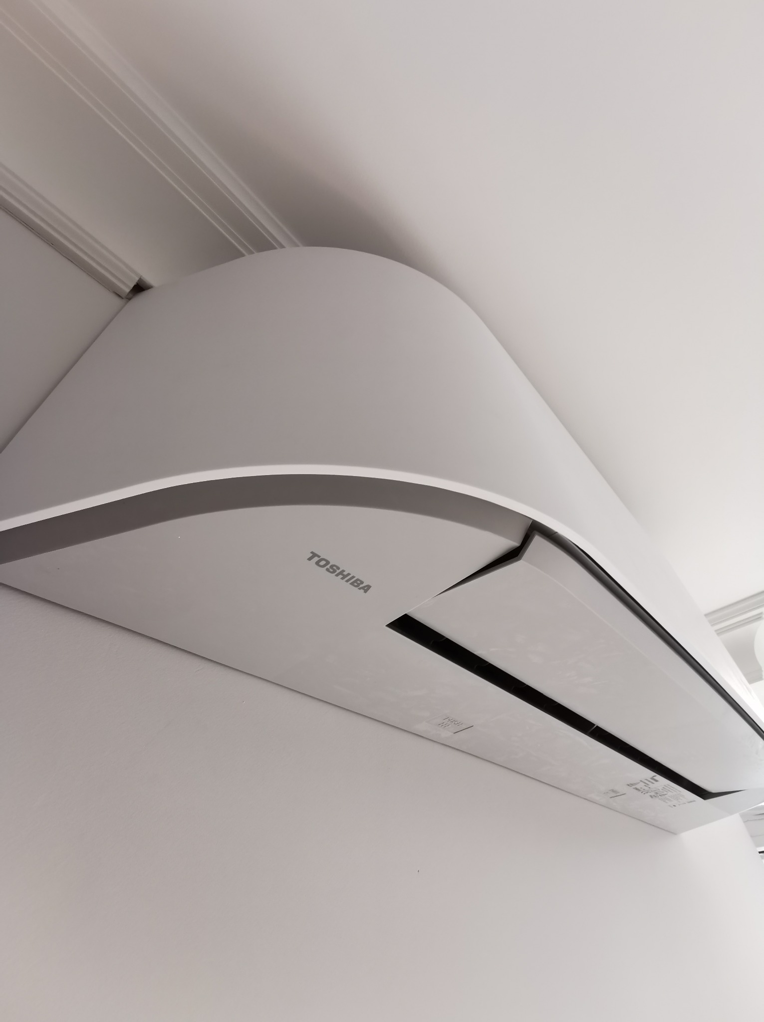 Biała jednostka wewnętrzna klimatyzacji Toshiba zamontowana na ścianie pod sufitem z ozdobną listwą.