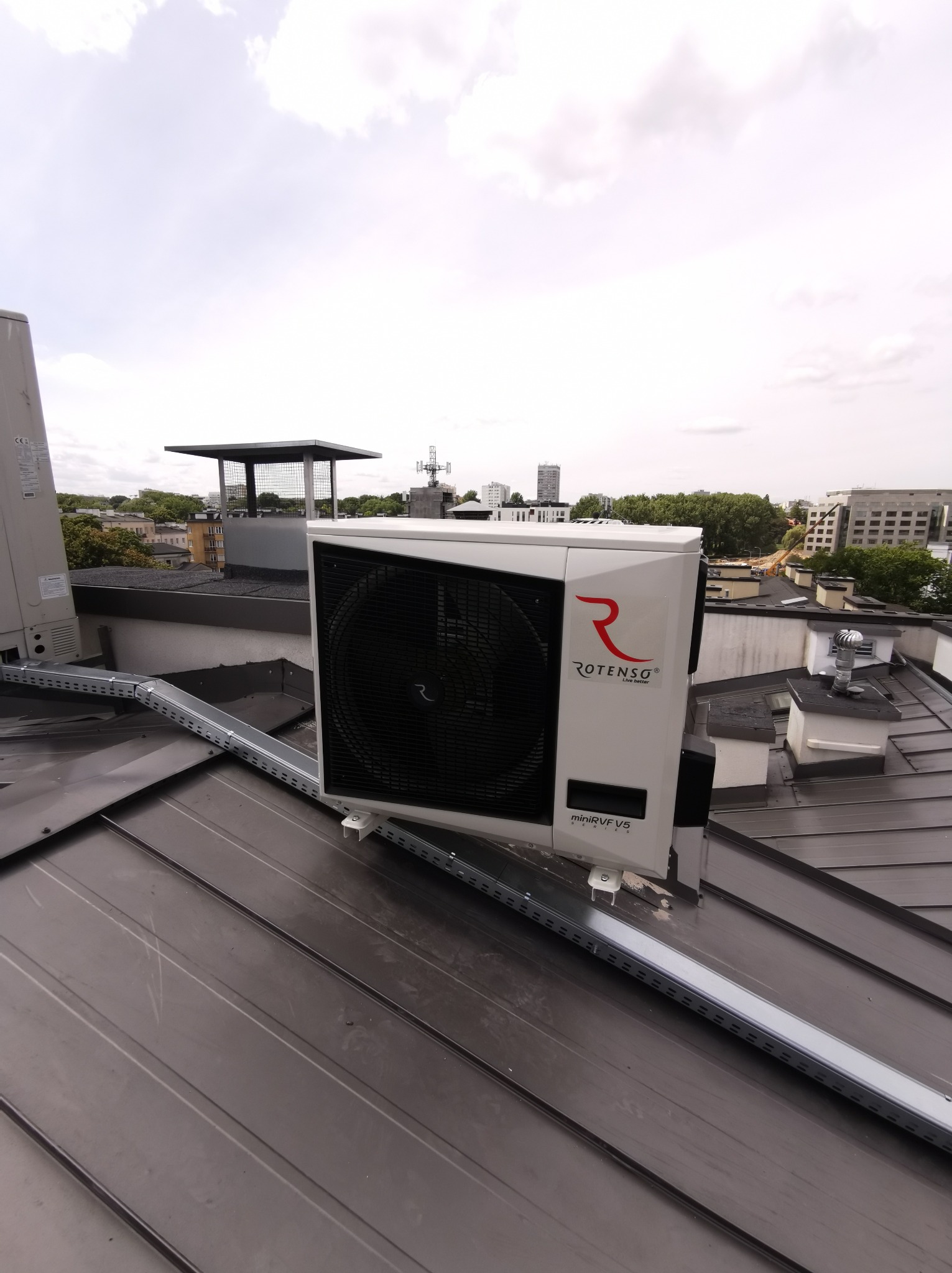 Zewnętrzna jednostka klimatyzacji Rotenso mini VRF V5 zamontowana na metalowym dachu budynku z widokiem na miejski krajobraz w tle.