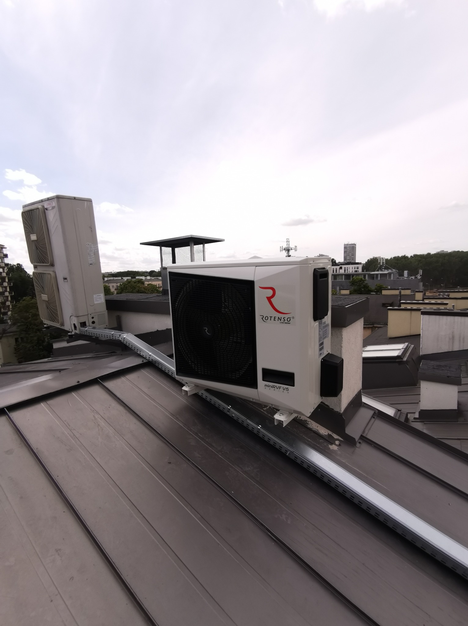 Dwie jednostki zewnętrzne klimatyzacji Rotenso miniVRF V5 zamontowane na metalowej konstrukcji na dachu budynku z blachy, widok na miasto w tle.