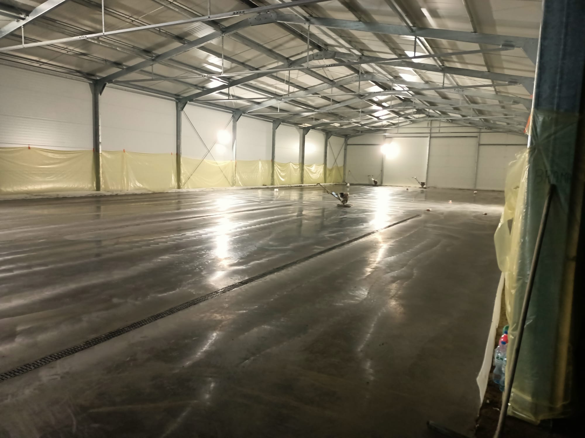 Wnętrze hali z wylewaną, mokrą posadzką przemysłową, zabezpieczone ściany folią, widoczne maszyny do polerowania.