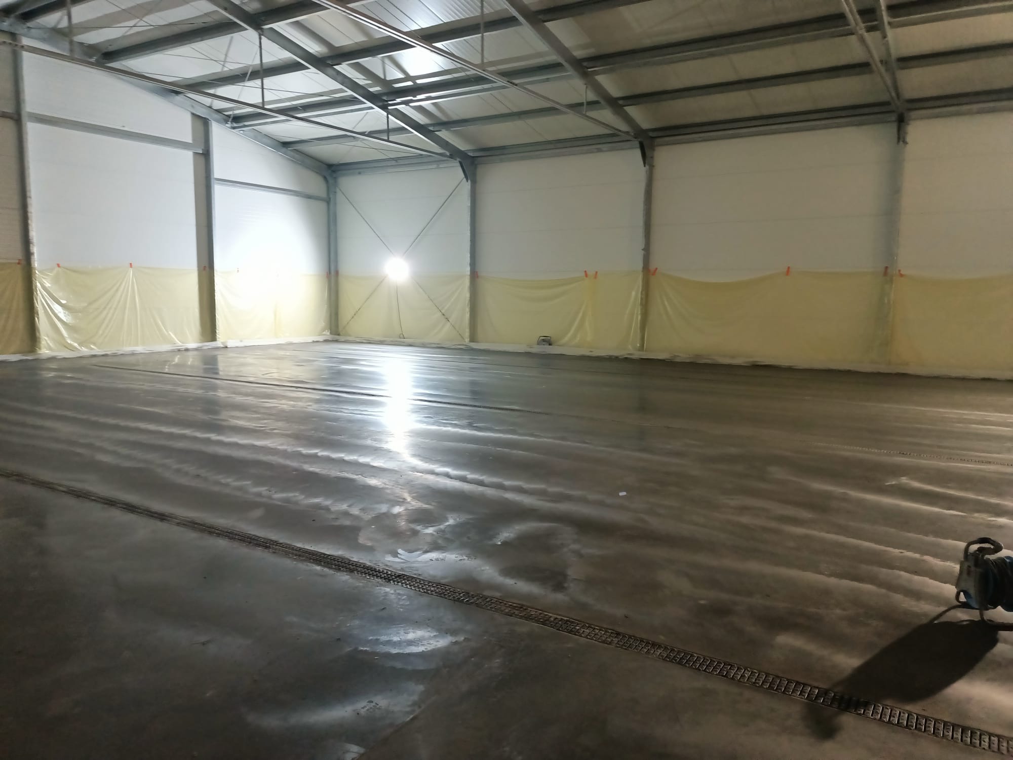Świeżo wylana, mokra posadzka w hali przemysłowej z widocznymi śladami po wylewce i kratką odpływową w podłodze, zabezpieczone ściany folią. Widoczny sprzęt do wylewania posadzek.