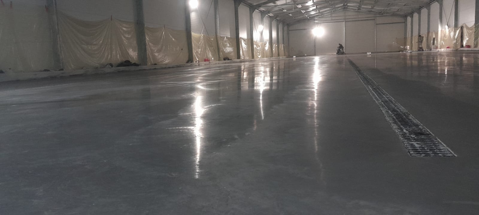 Gładka, szara posadzka w hali przemysłowej z widocznym odpływem liniowym i pracownikiem obsługującym maszynę do polerowania w tle.