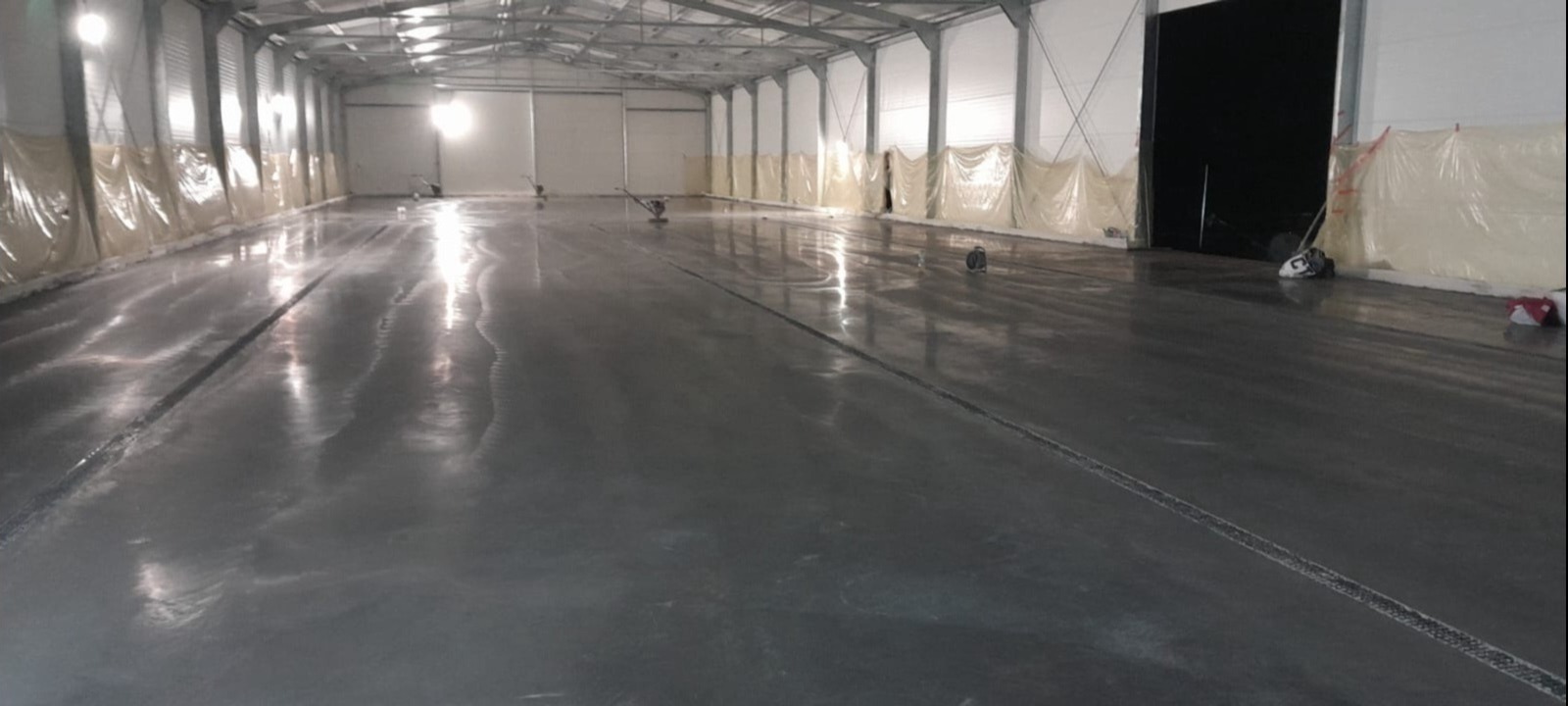 Błyszcząca, szara posadzka w hali przemysłowej, zabezpieczona folią przy ścianach, z widocznymi maszynami do polerowania.