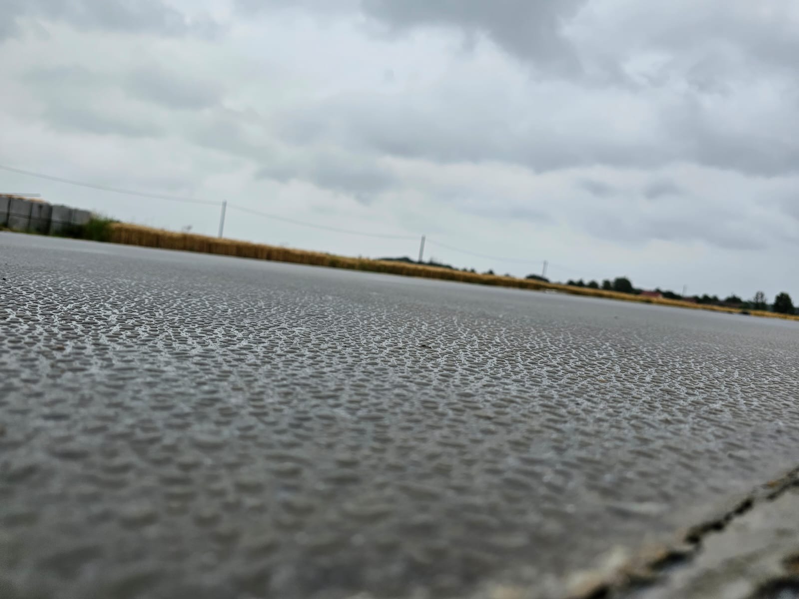 Szara, mokra powierzchnia z bliska, prawdopodobnie betonowa, z widoczną fakturą i odbiciami, w tle pole i pochmurne niebo.