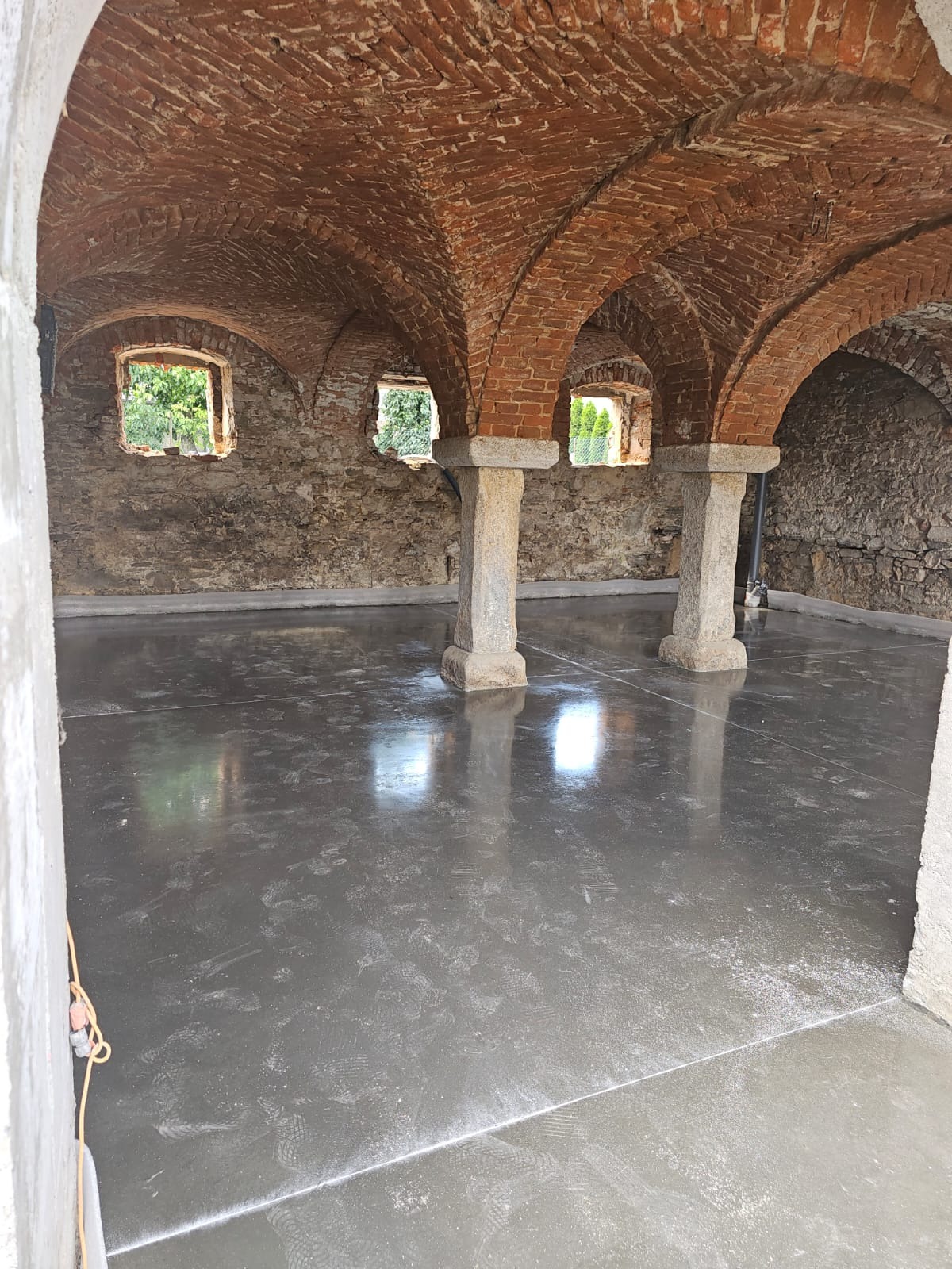 Świeżo wylana, gładka posadzka w zabytkowym pomieszczeniu z ceglanym sklepieniem łukowym i kamiennymi filarami, widoczne ślady po butach.
