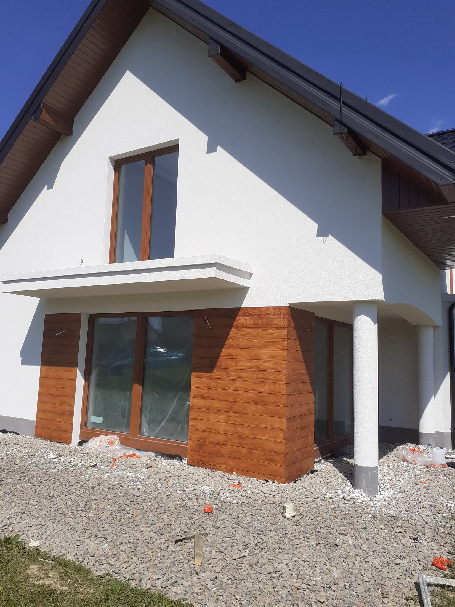 Nowoczesny dom jednorodzinny z białą elewacją i drewnianymi panelami dekoracyjnymi wokół okien, teren wokół domu wysypany kamieniami, okna zabezpieczone folią, widoczne elementy rusztowania.
