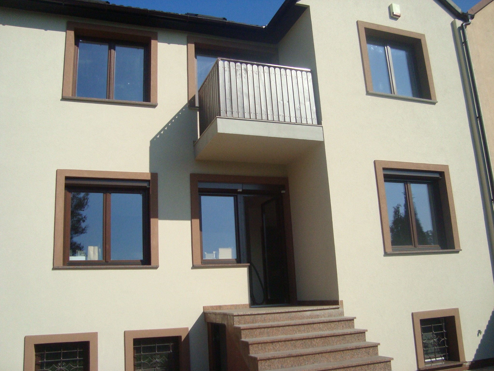 Jasna elewacja budynku z brązowymi obramowaniami okien i balkonem z drewnianą balustradą, widoczne schody wejściowe z brązowego granitu.